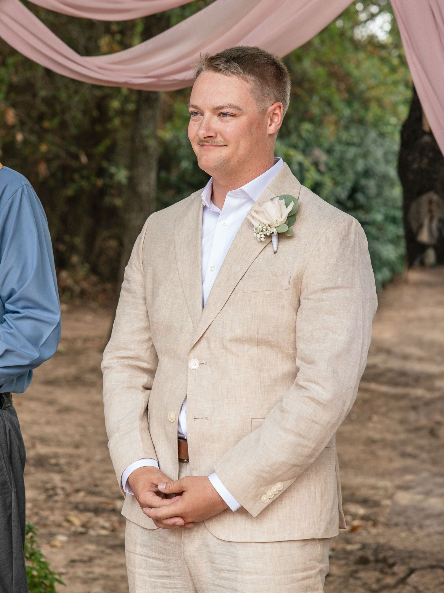A smiling groom in a beige linen suit with a floral boutonniere stands with clasped hands at an outdoor wedding ceremony.