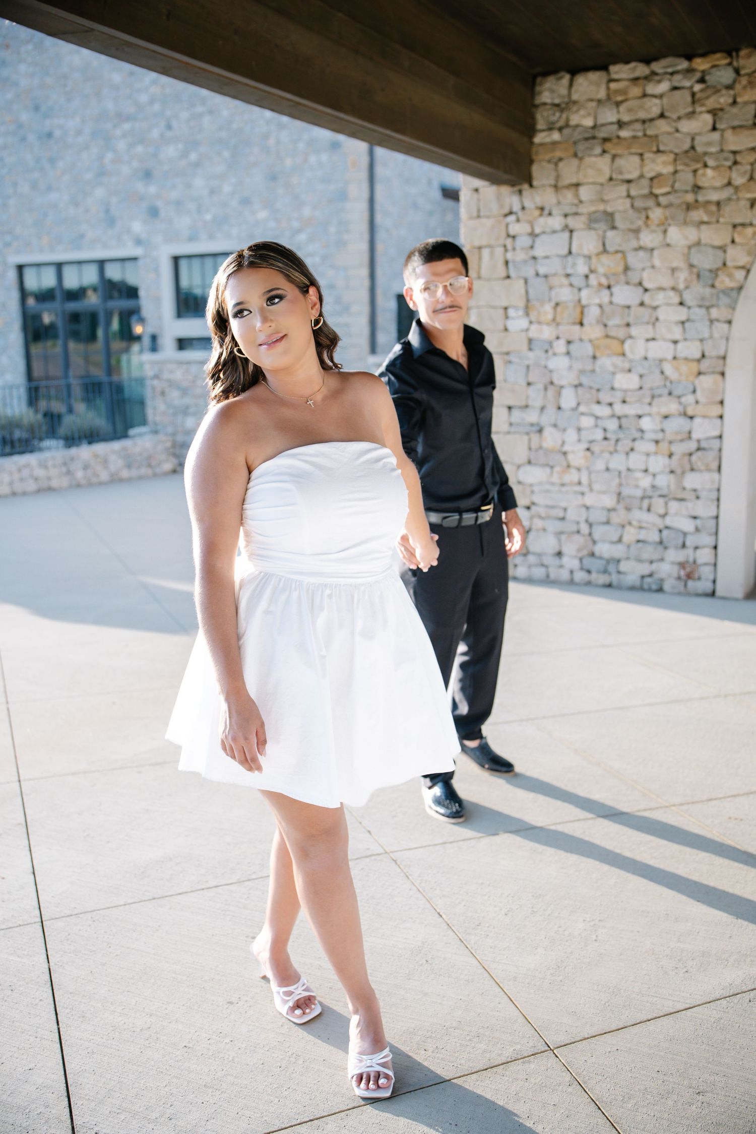 A woman in a white strapless mini dress walks confidently outdoors while a man in black follows behind near a stone wall.