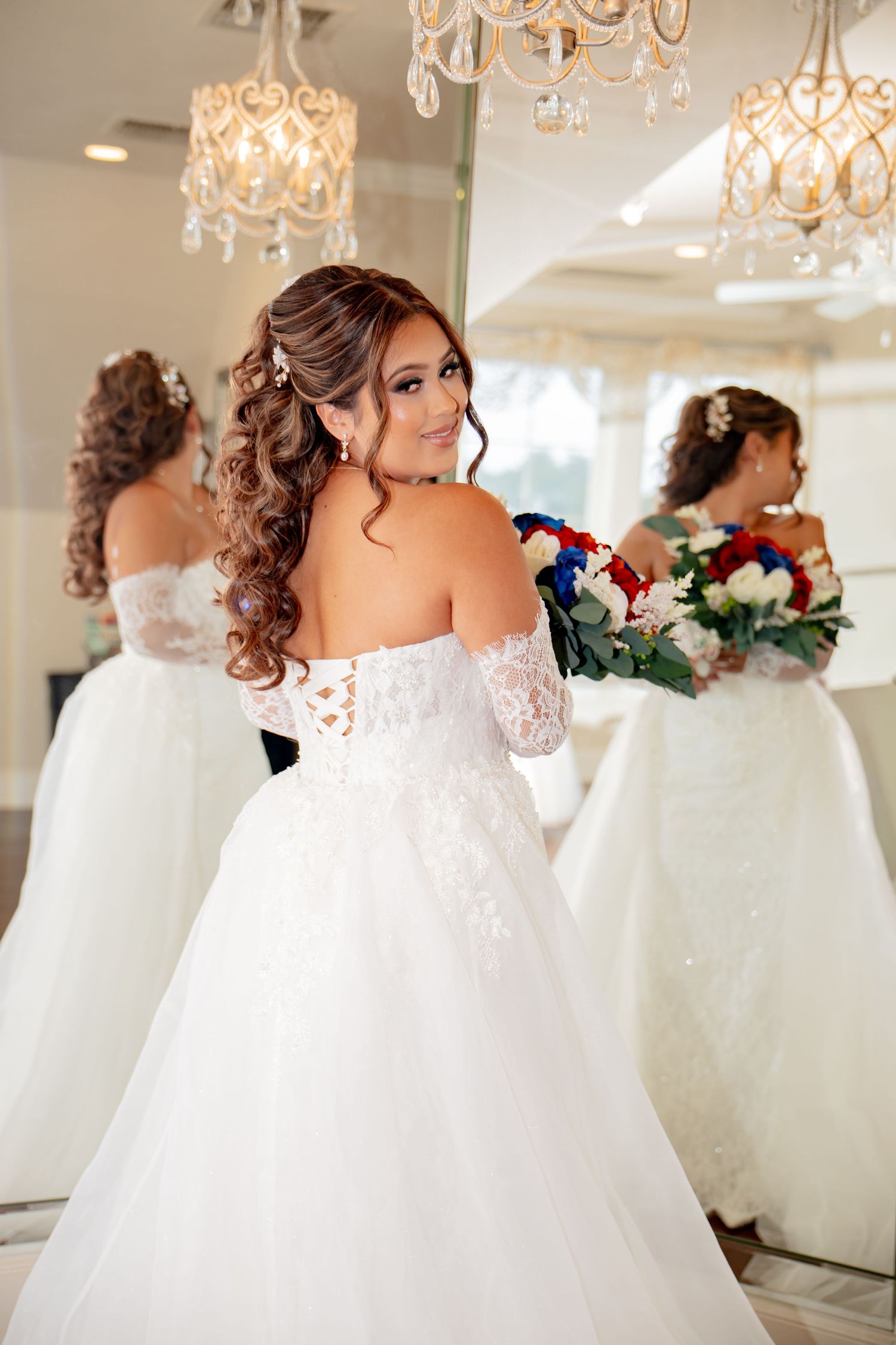 Bride in off-shoulder lace wedding gown holds bouquet while posing in front of mirror.