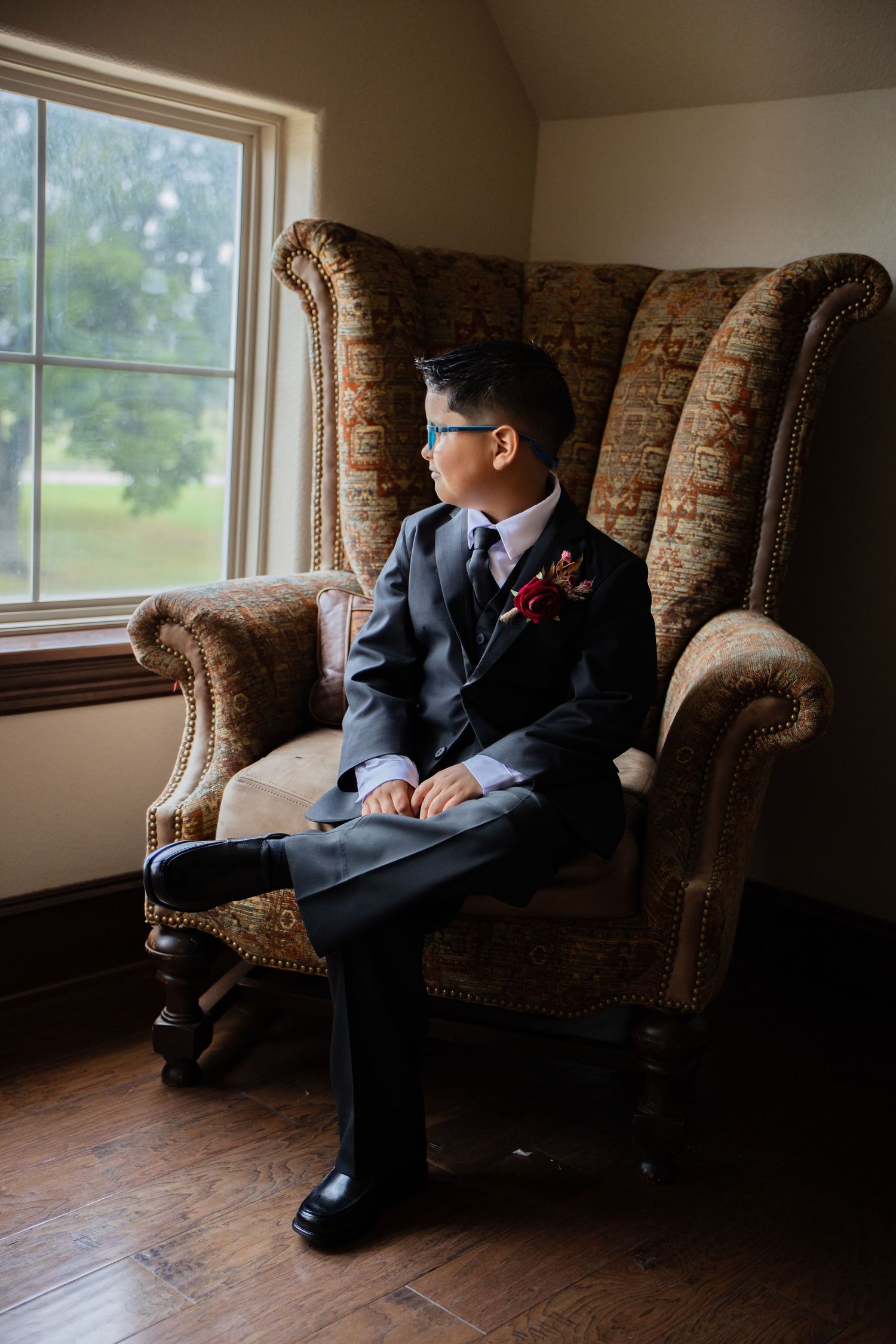 Young ring bearer in dark suit with red boutonniere sits in ornate wingback chair gazing out window.