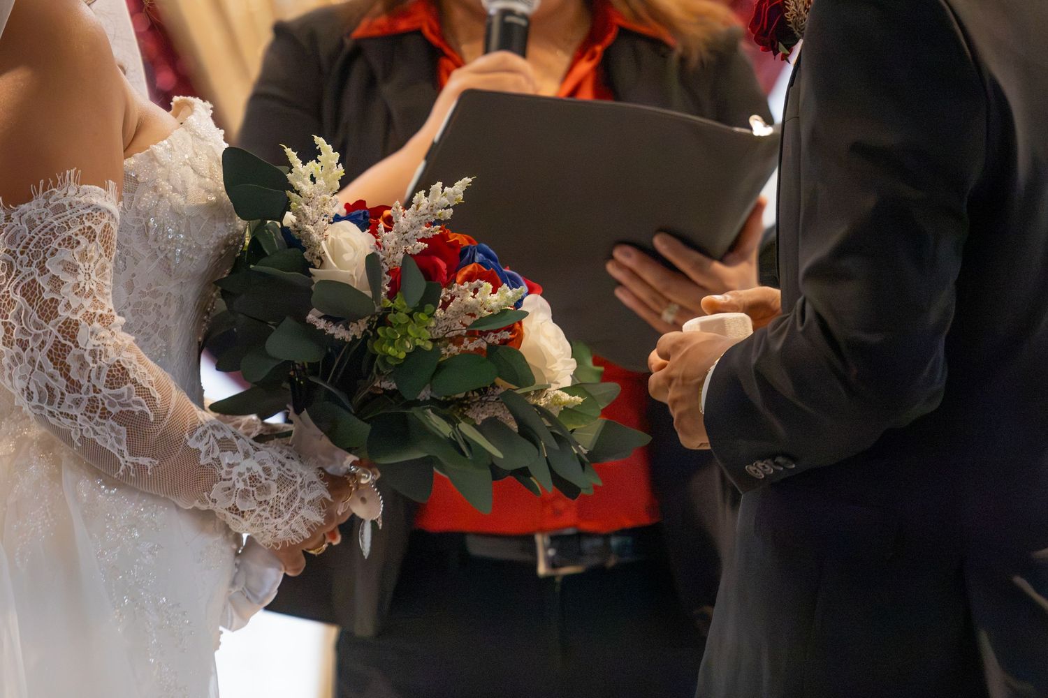 Bride holding a bouquet of red and white flowers exchanges vows with groom as officiant reads from a binder.