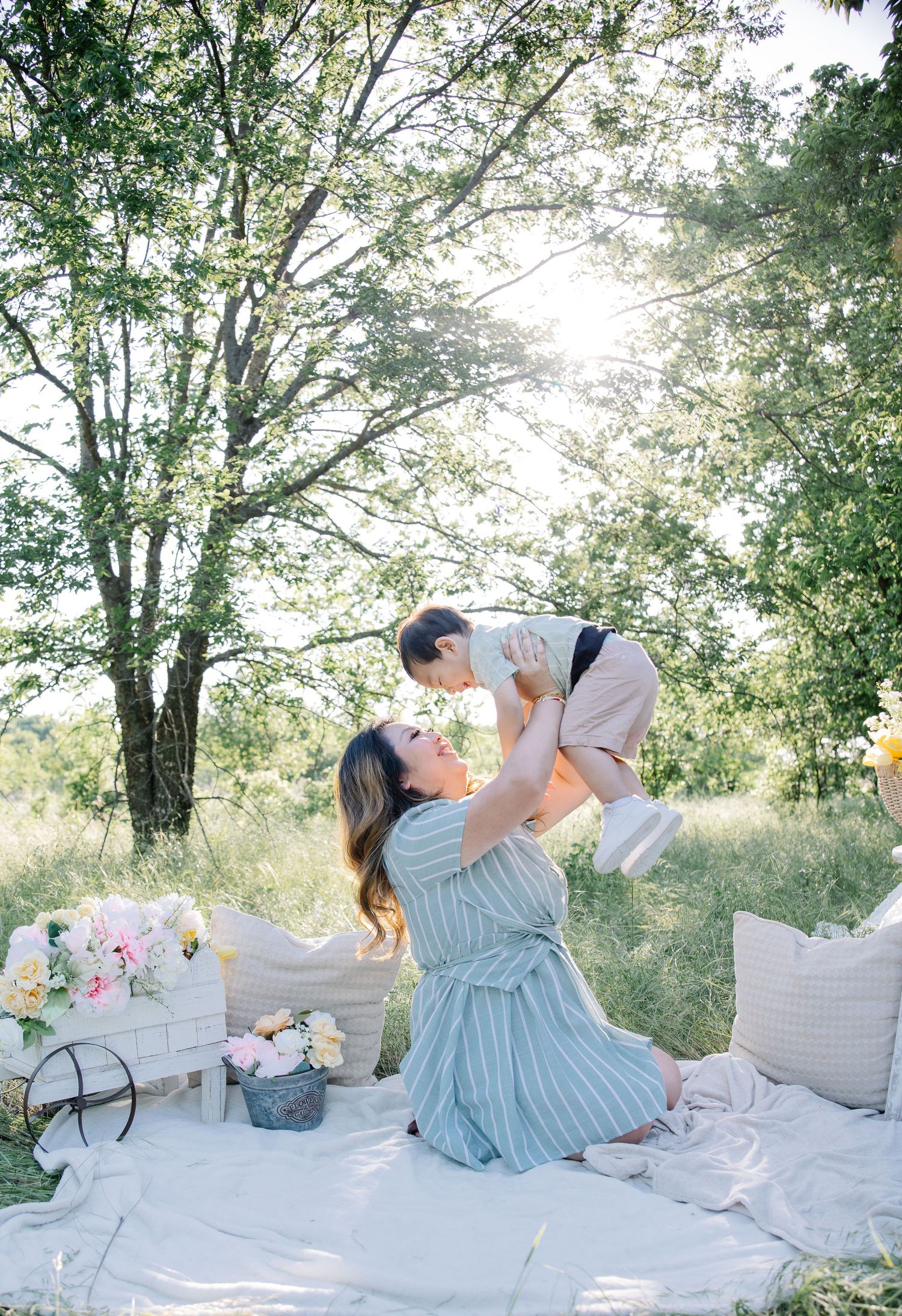 A mother kneels on a blanket outdoors, lifting her toddler up to her face for a kiss in a sunlit meadow.