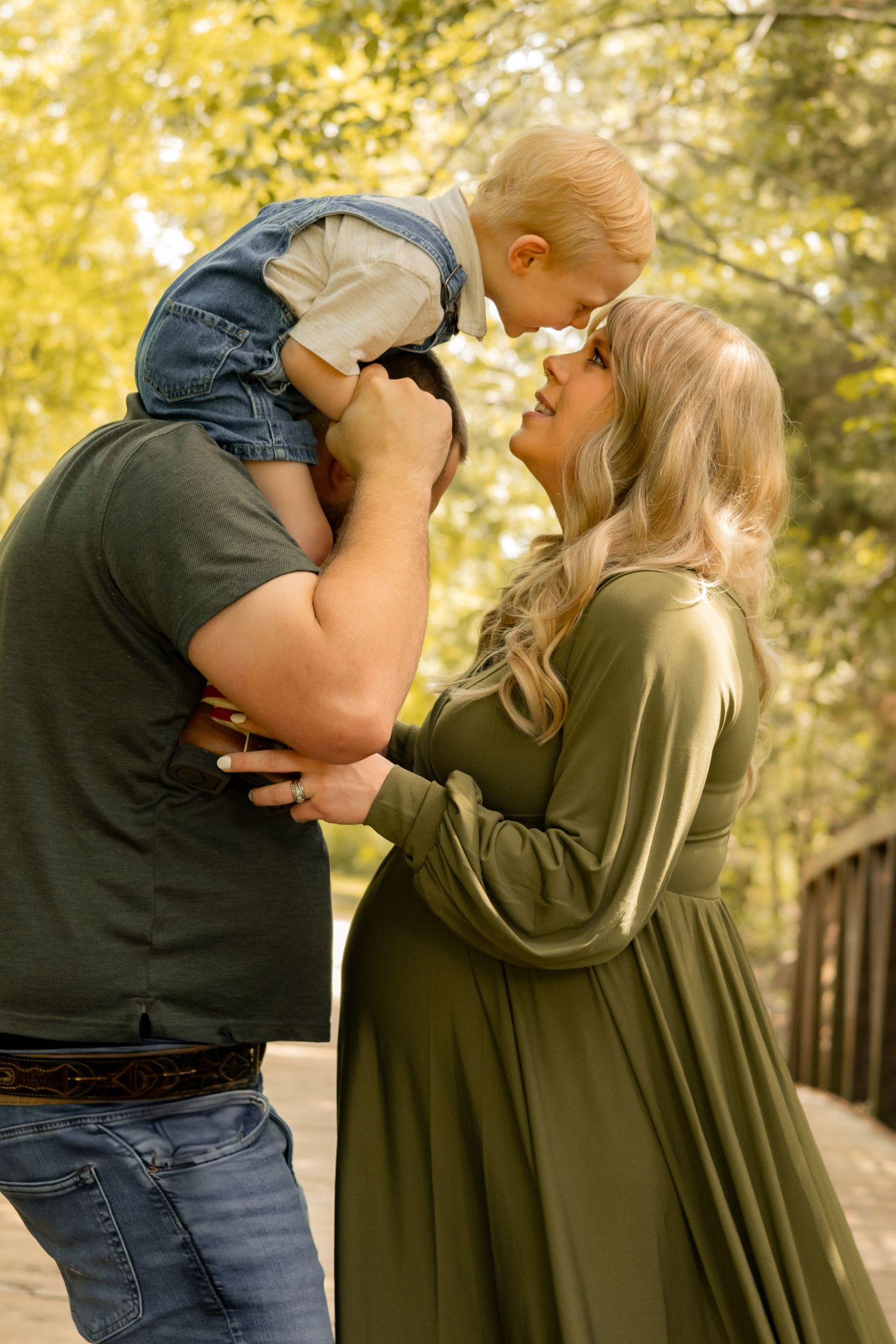 Parents in olive and dark green outfits lift laughing toddler between them during outdoor maternity photo session.