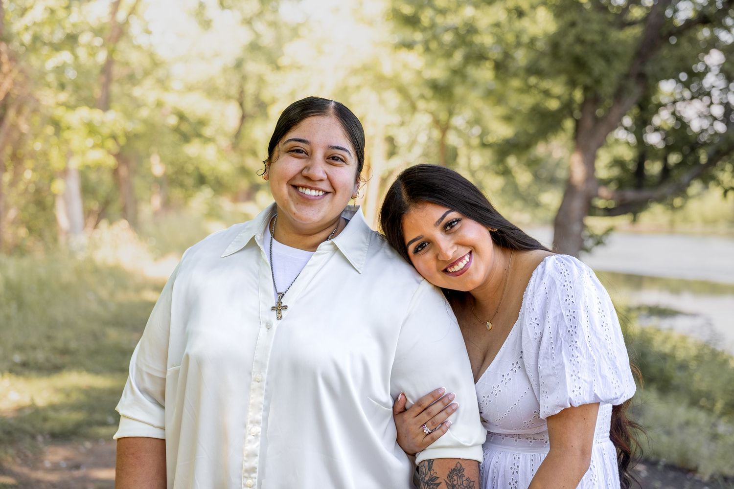 Two smiling women posing closely together outdoors with soft natural bokeh background and warm sunlight.