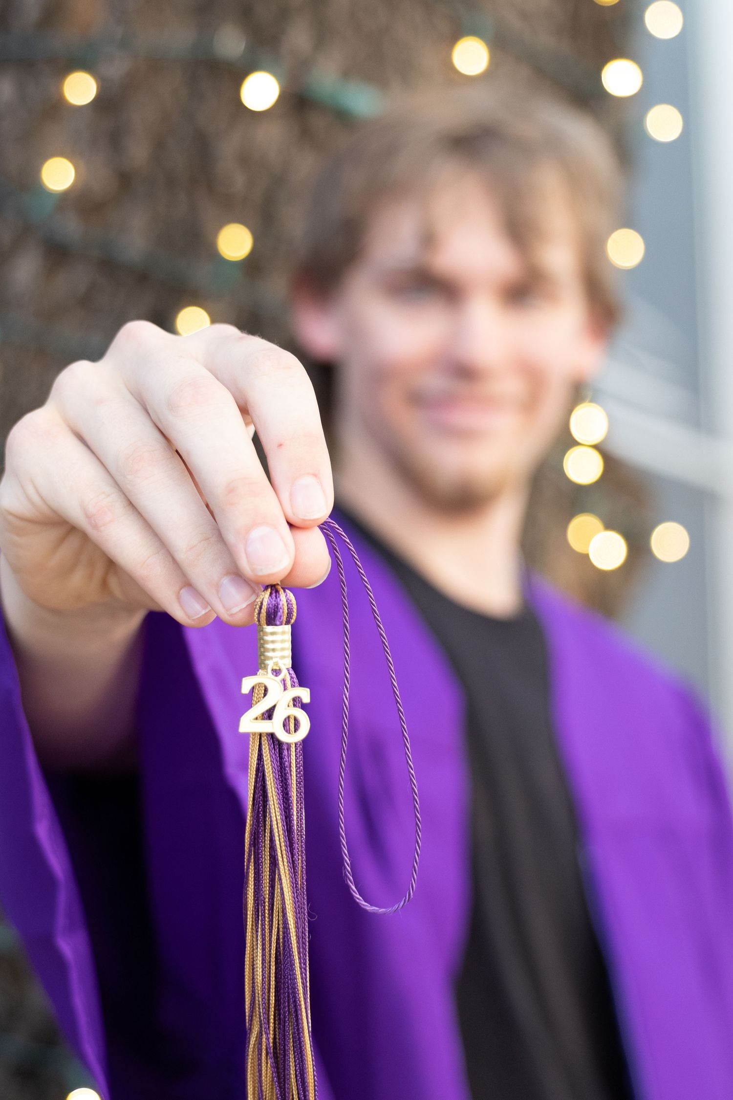 Smiling graduate in purple gown holding a tassel with a 26 charm against bokeh lights background.