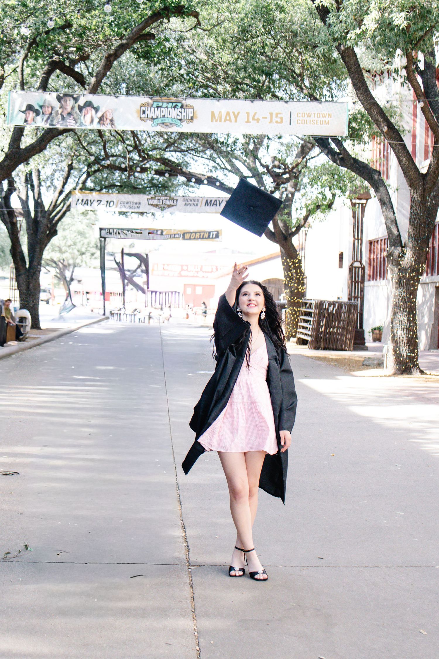 Young woman in pink dress and graduation gown tosses cap on tree-lined street.