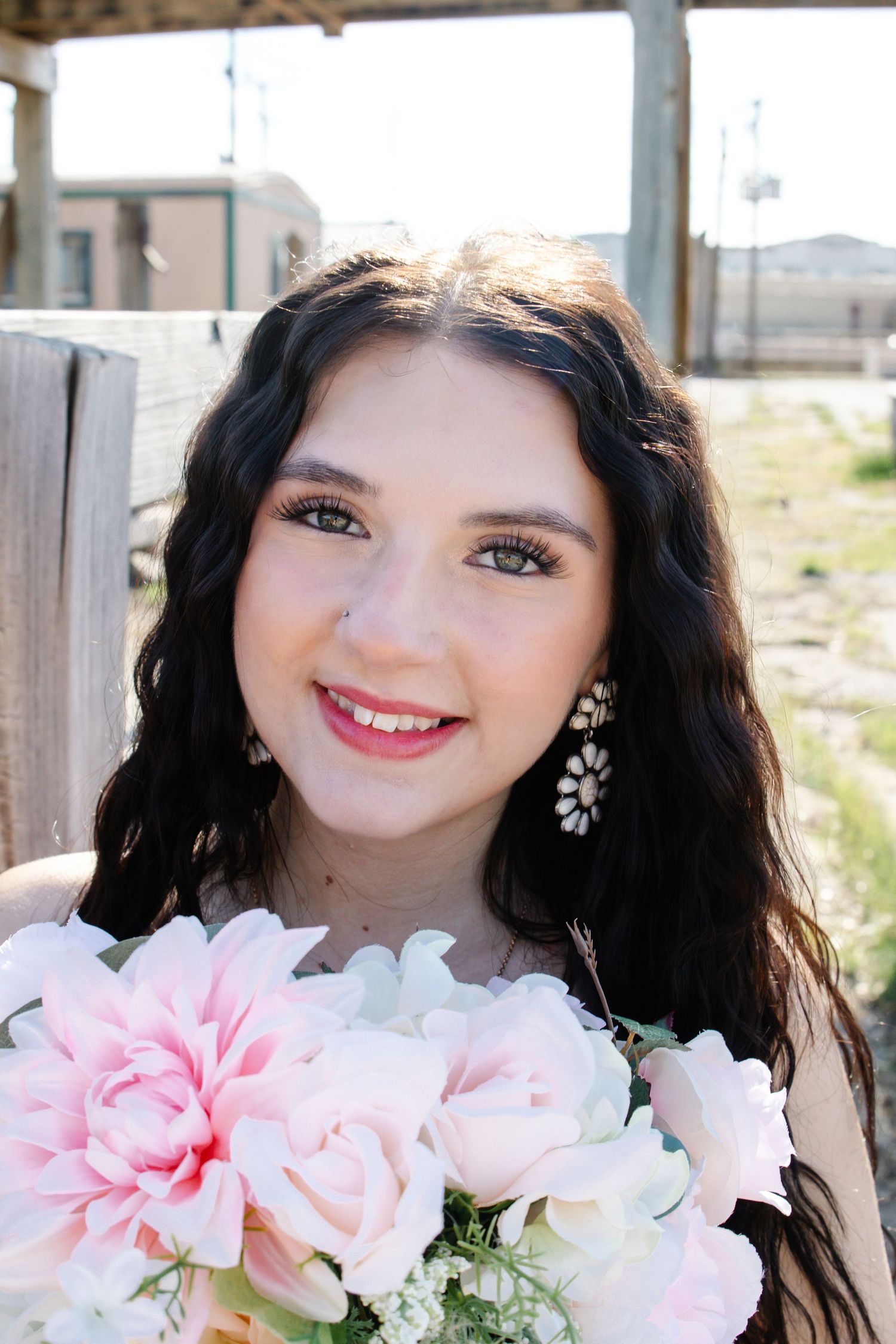 Smiling young woman with dark curly hair holding pink roses wearing floral earrings outdoors.