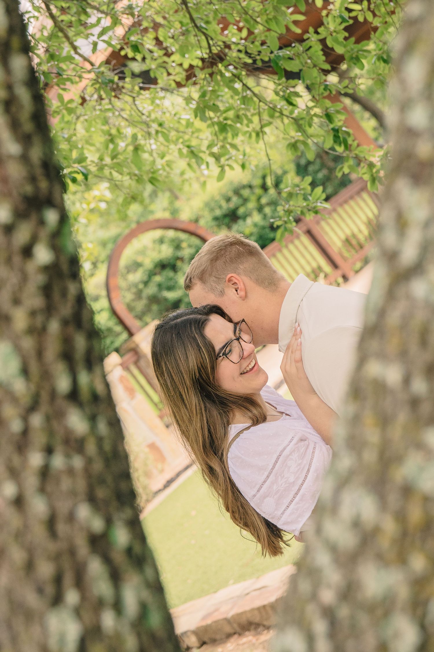 Couple sharing a tender moment framed by tree trunks in a beautiful outdoor garden setting.