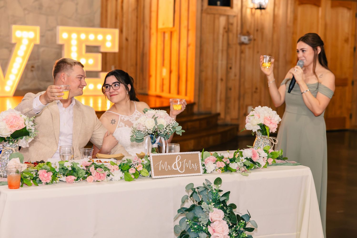 Bridesmaid gives a toast with a microphone as the newlyweds raise glasses at a floral-decorated head table in a rustic barn.