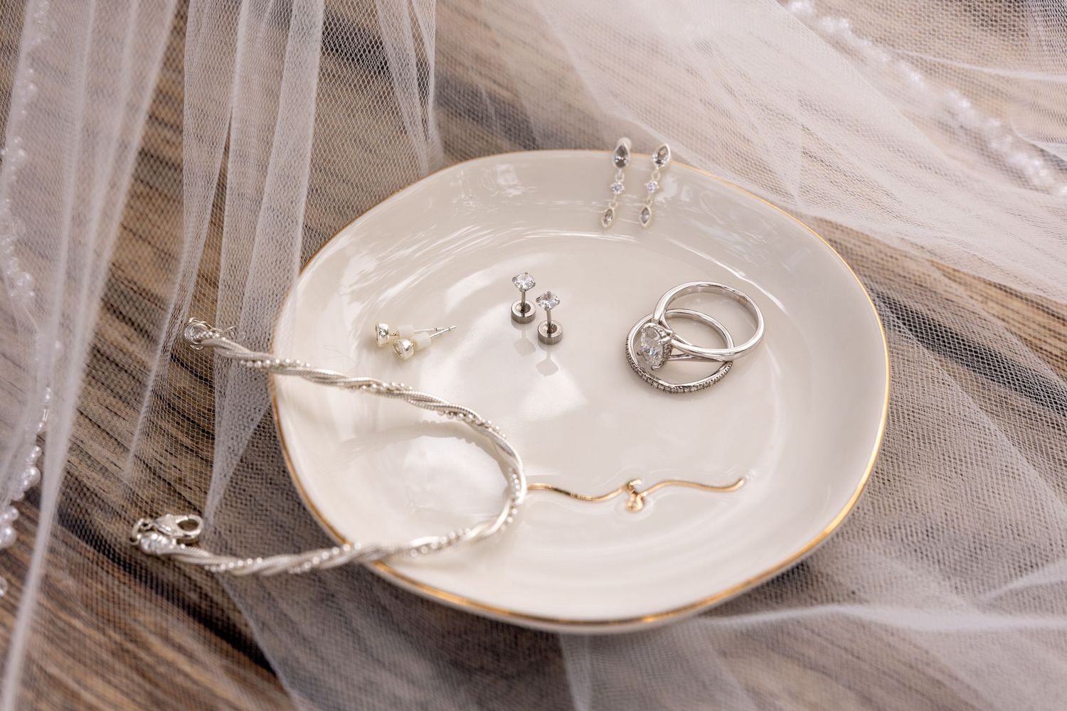 White ceramic dish with gold rim holding silver rings, stud earrings, and delicate bracelets on a rustic wood table beside sheer tulle veil.