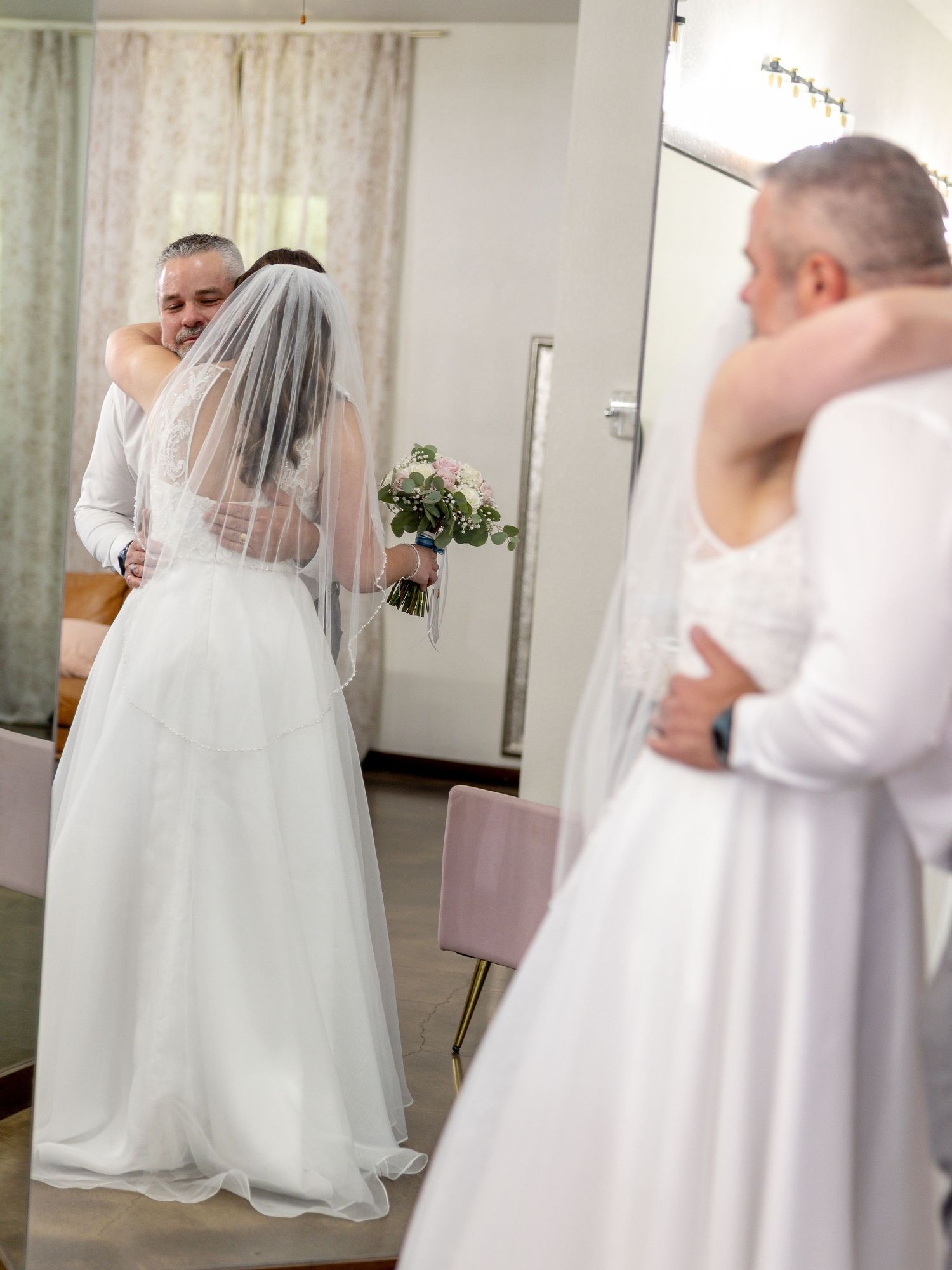 A bride in a white gown and veil embraces a man in a white shirt, with the moment reflected in mirrors, as she holds a floral bouquet.