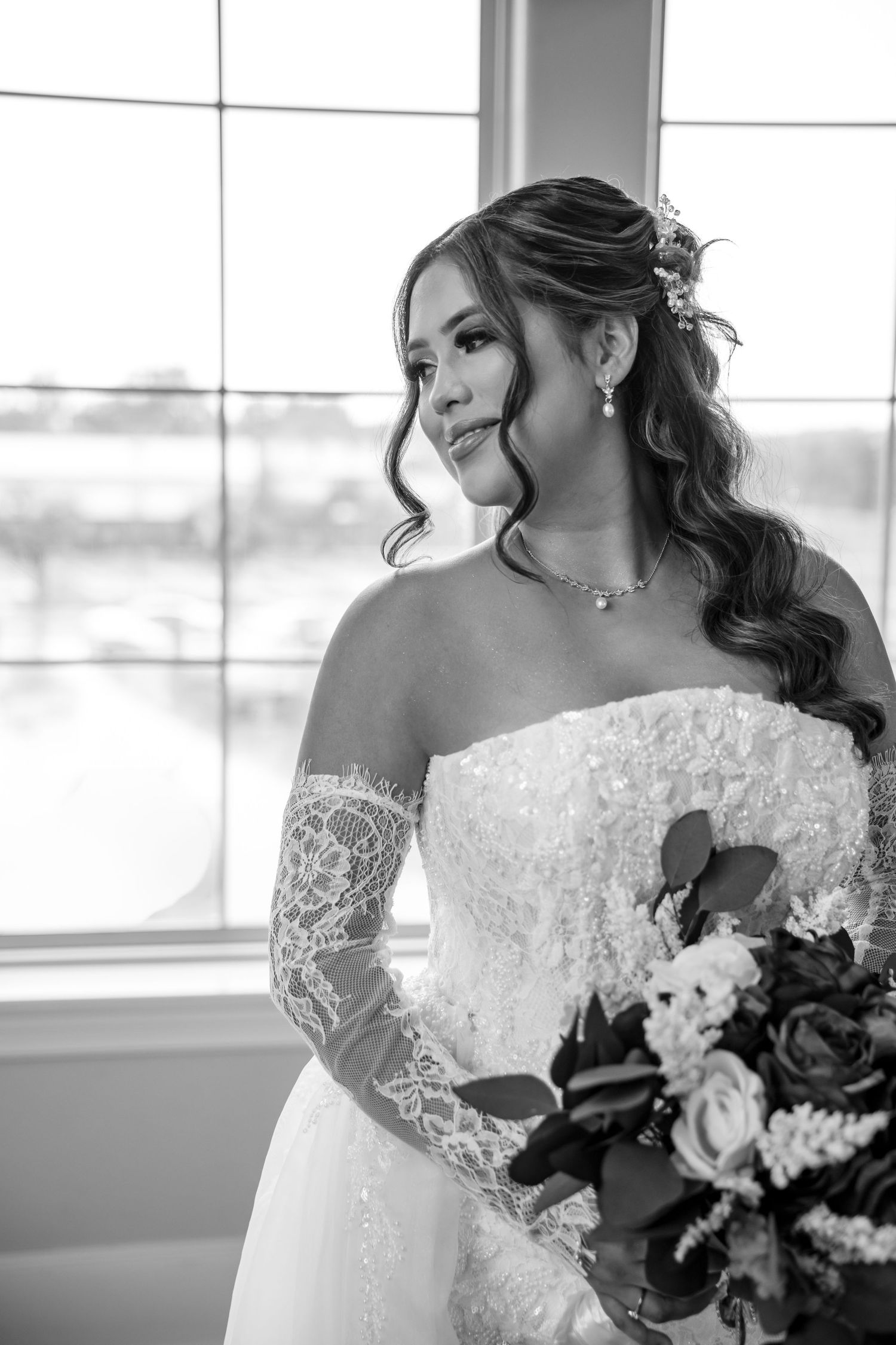 Black and white of a bride in a strapless lace wedding gown and lace gloves, holding a rose bouquet beside a bright window. Burleson Texas
