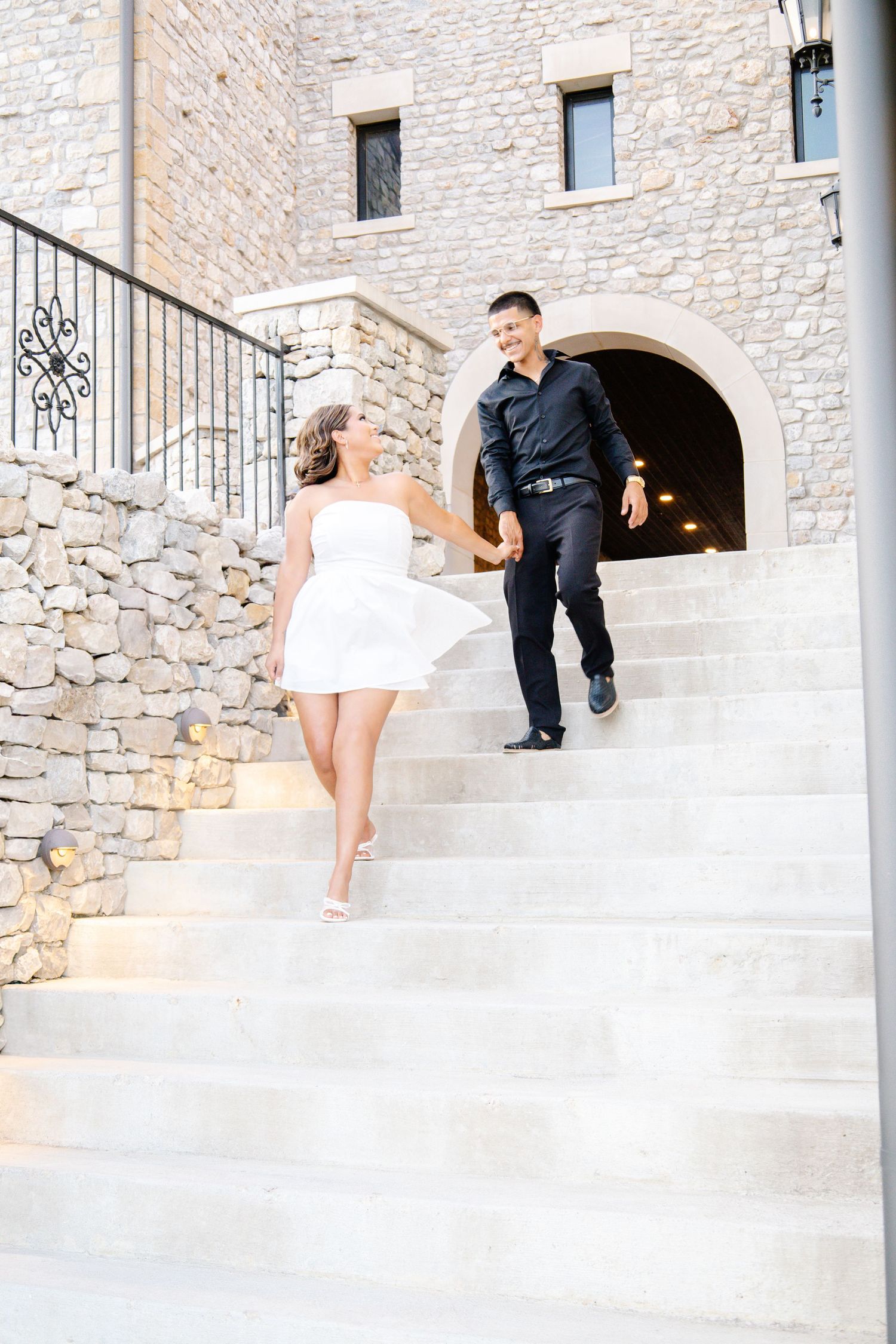 Couple in black and white outfits holding hands while descending stone stairs outside a rustic building.