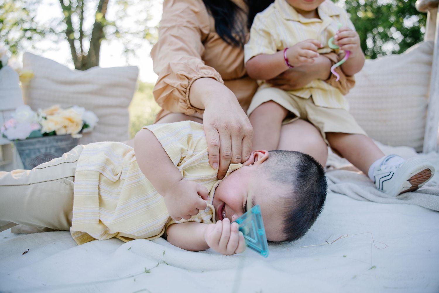 A mother in yellow sits outdoors with two young children dressed in matching outfits, one baby lying down with a pacifier.