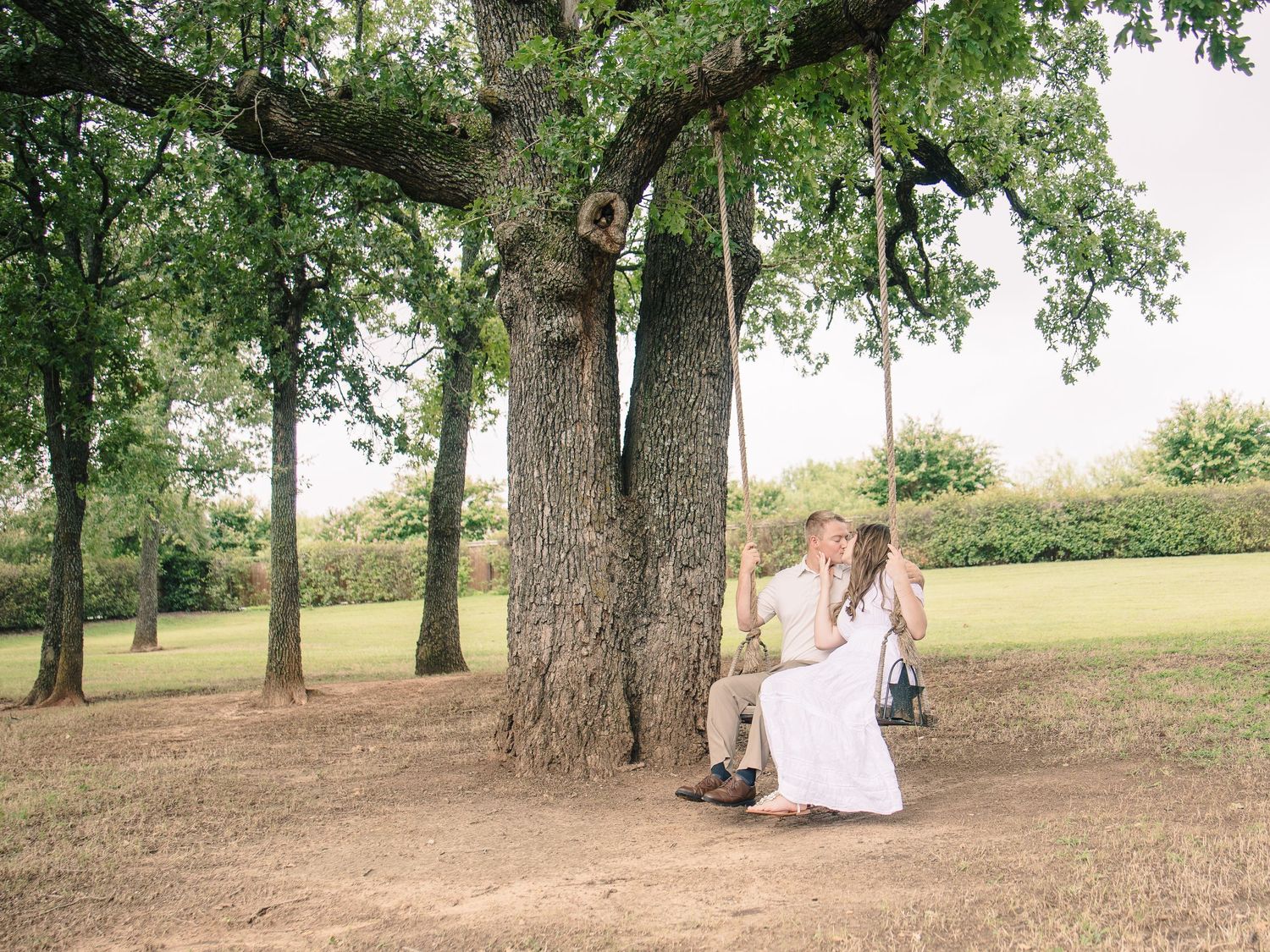 Couple sharing a romantic kiss on a tree swing in a lush green park, dressed in wedding attire.