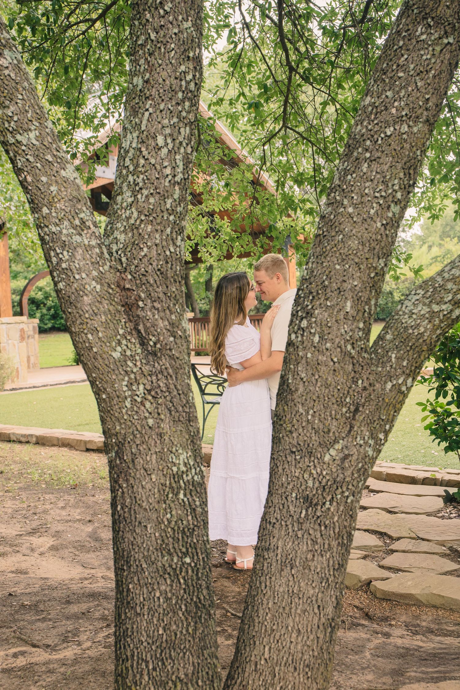 Couple in white embracing between tree trunks in a lush green garden setting.