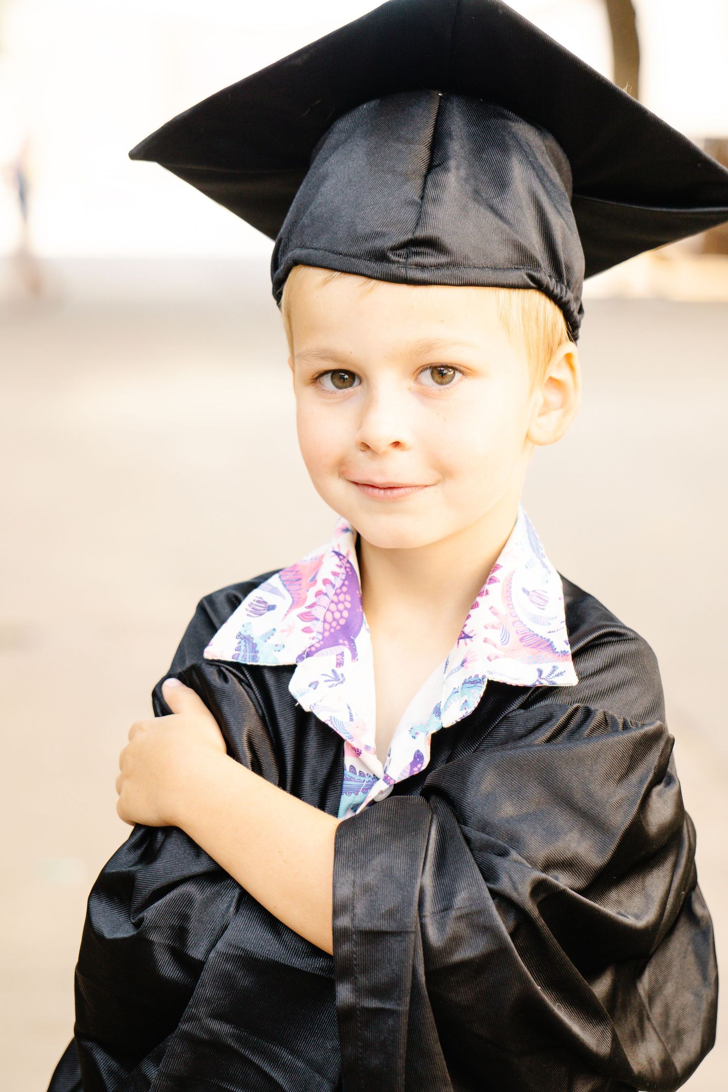 Cute young blonde boy in black graduation gown and cap with arms crossed smiling.