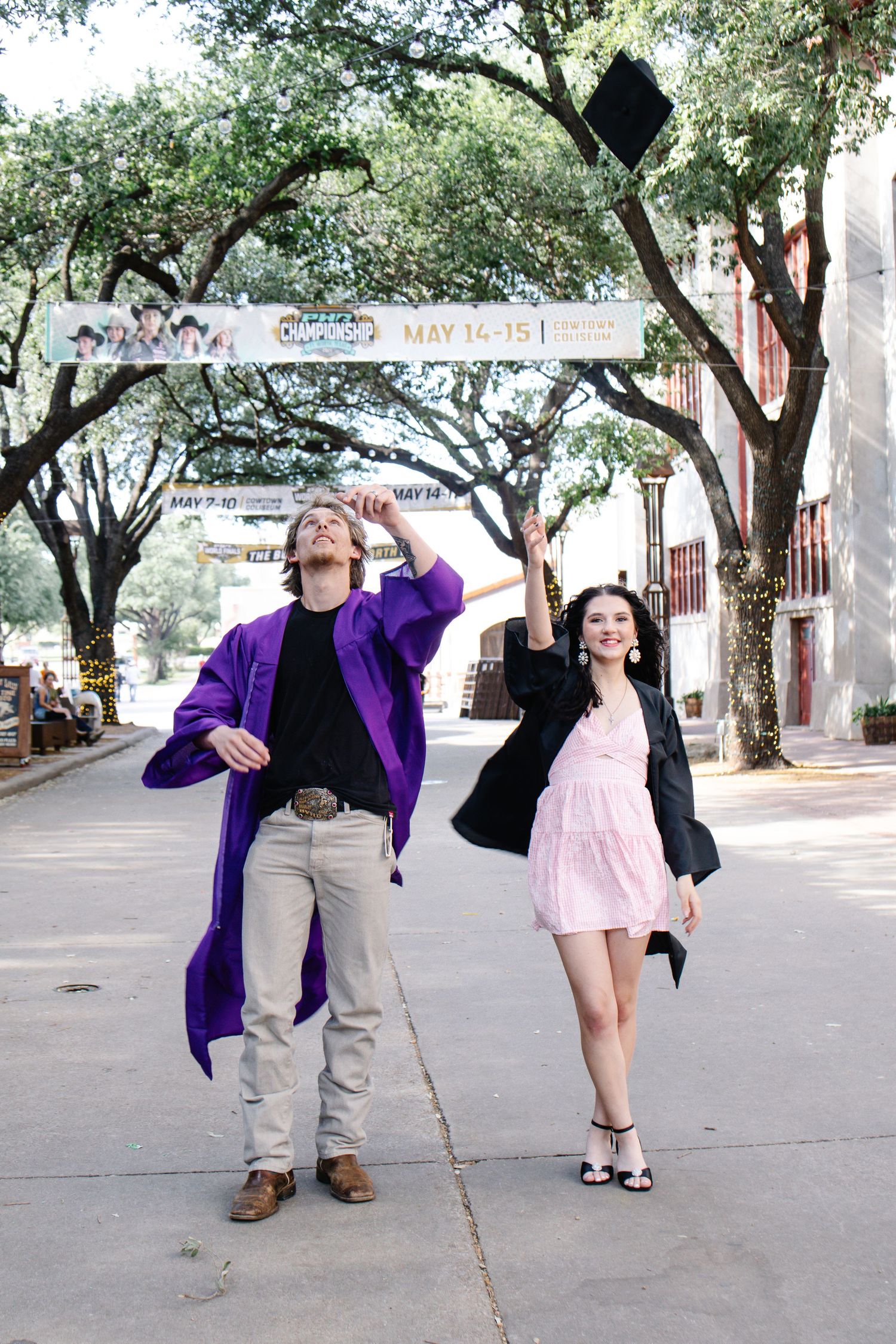 Two graduates celebrating by tossing caps on a scenic tree-lined outdoor street.