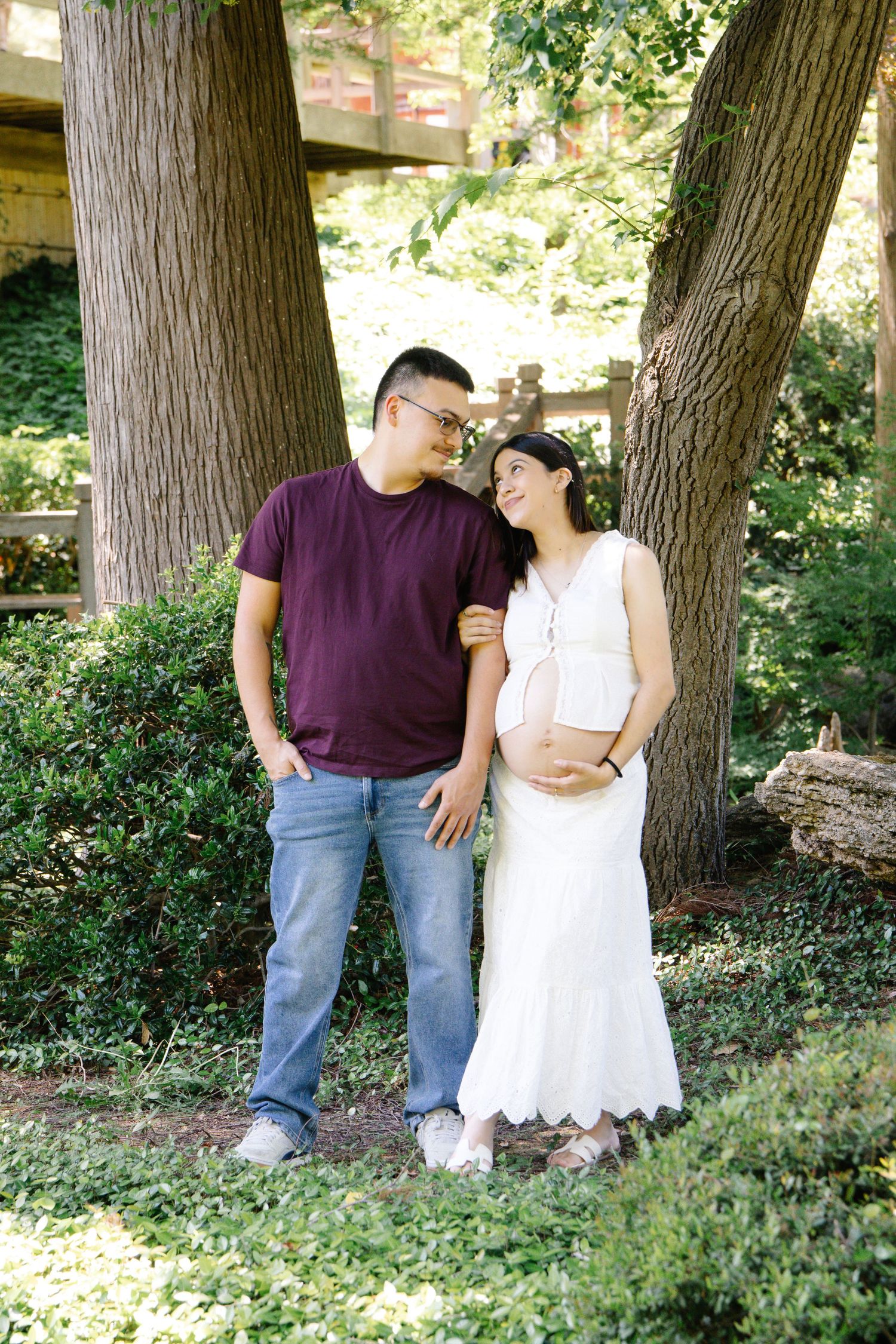 Smiling pregnant couple sharing a loving glance while standing among trees in a garden setting.