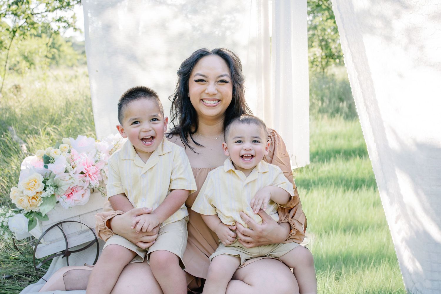 A smiling mother sits outdoors with her two young sons dressed in matching yellow outfits, surrounded by pink flowers.