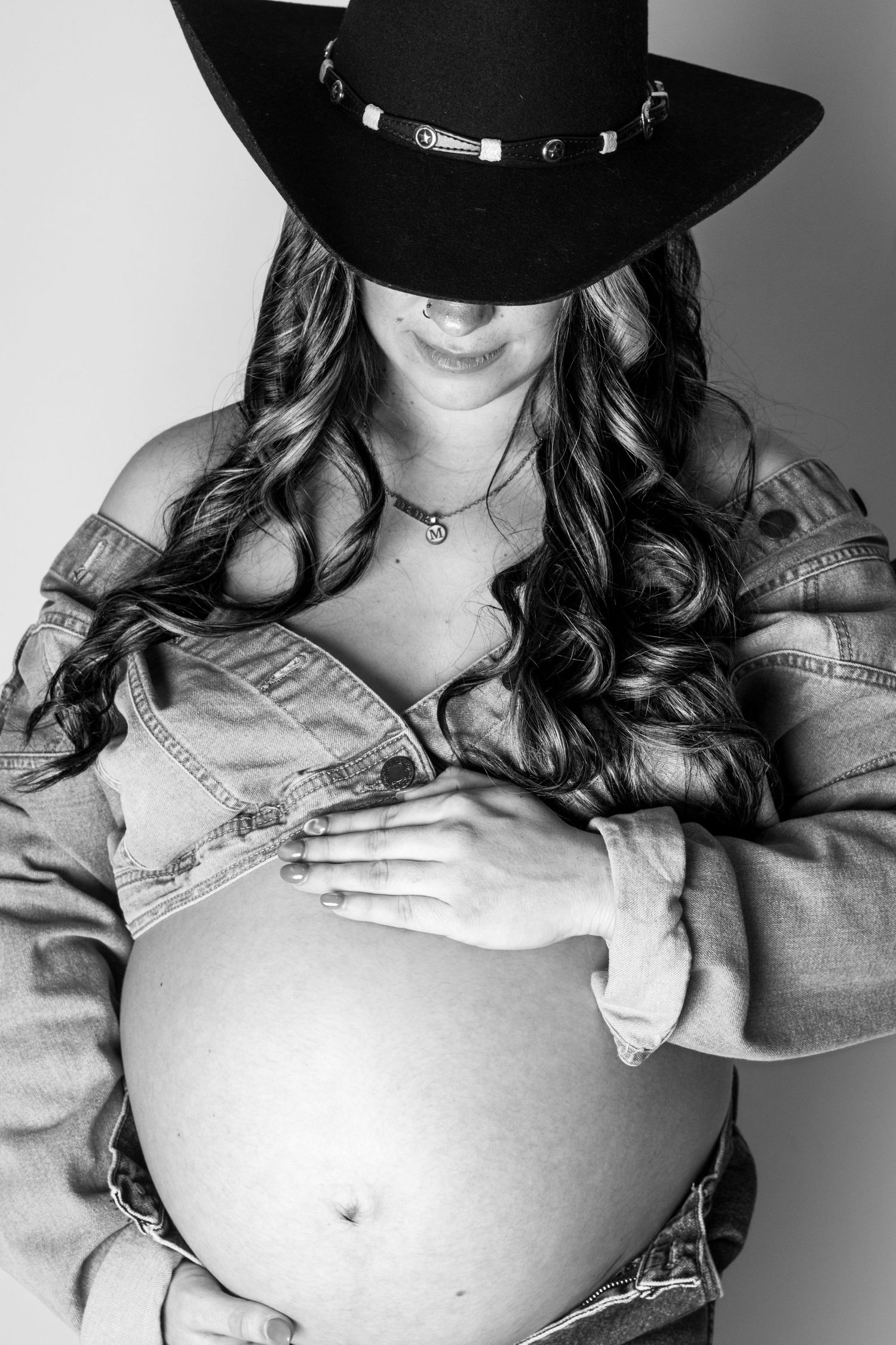 Black and white photo of a pregnant woman in a cowboy hat and denim jacket cradling her baby bump.