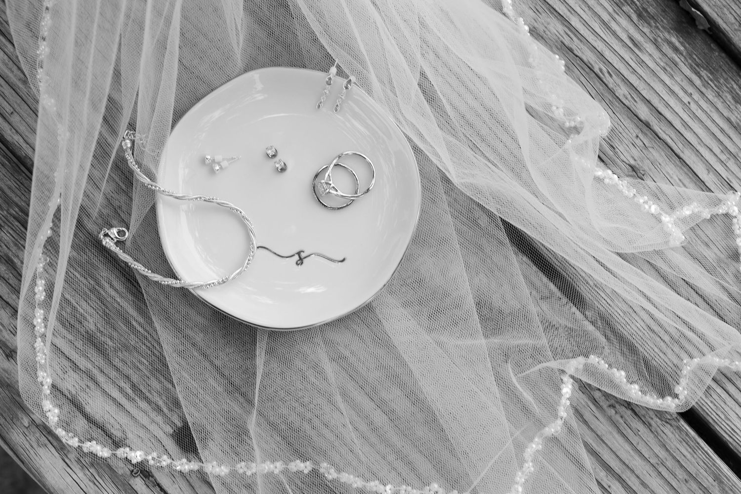 Black and white photo of wedding rings, earrings and a bracelet in a small dish resting on a wooden table beneath a beaded bridal veil.