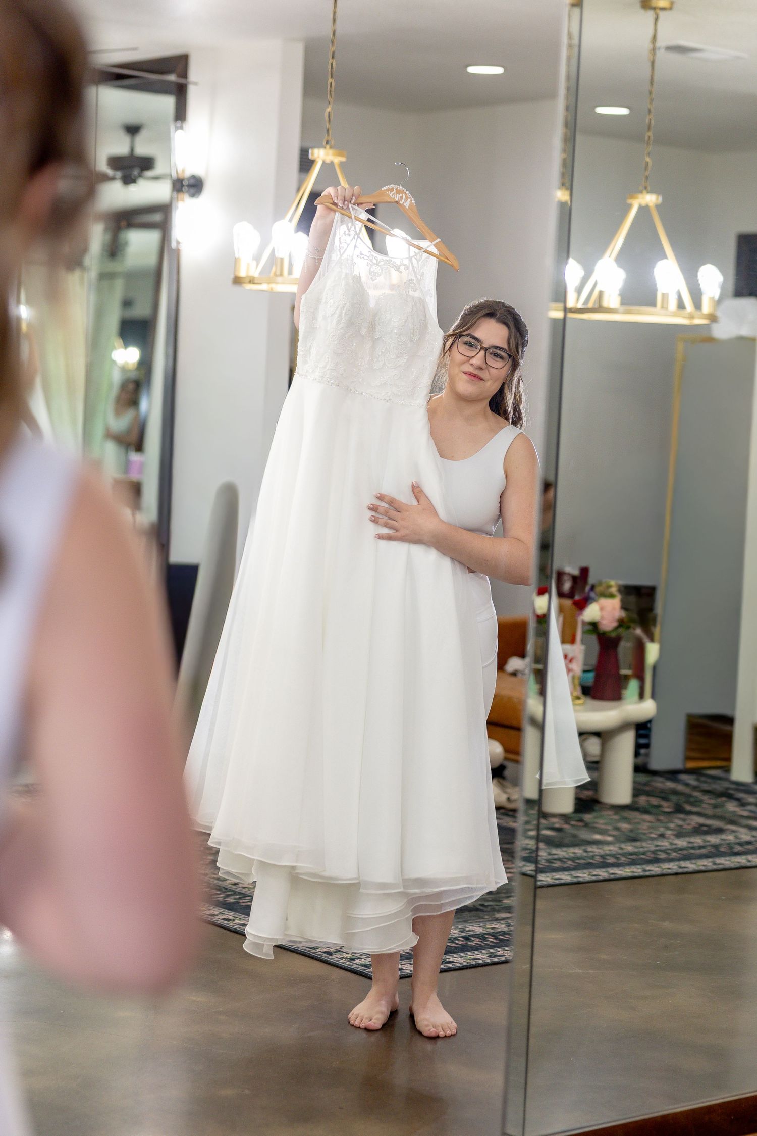 Bride in glasses stands barefoot in a bridal suite, holding her white wedding dress on a hanger and smiling at her mirror reflection.