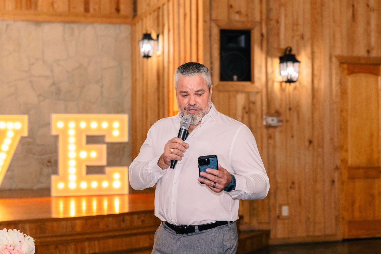 Man in a white shirt speaks into a microphone while holding a phone at a rustic wedding reception with marquee letters.