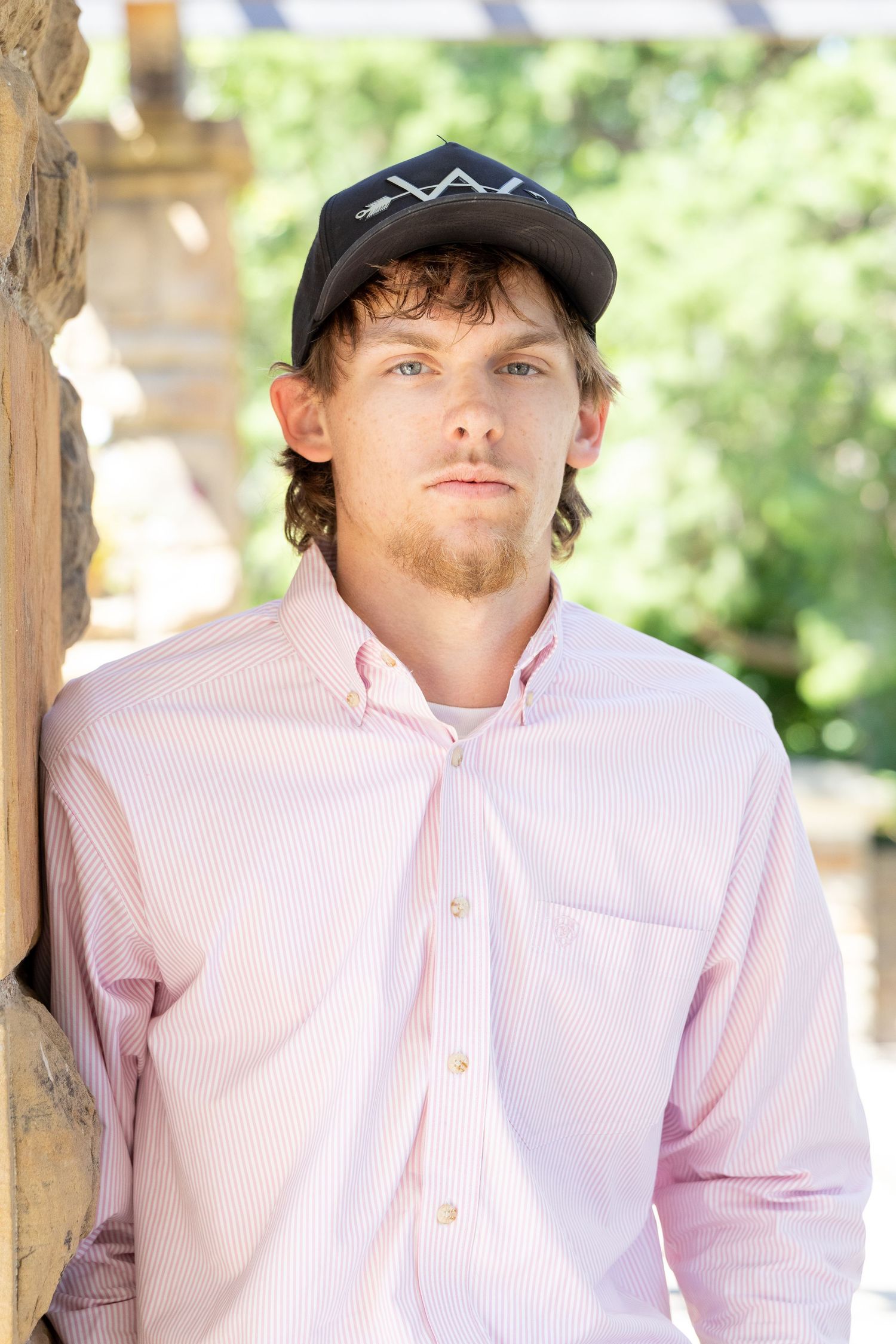 Young man with beard in pink striped shirt and black cap leans against stone wall outdoors.