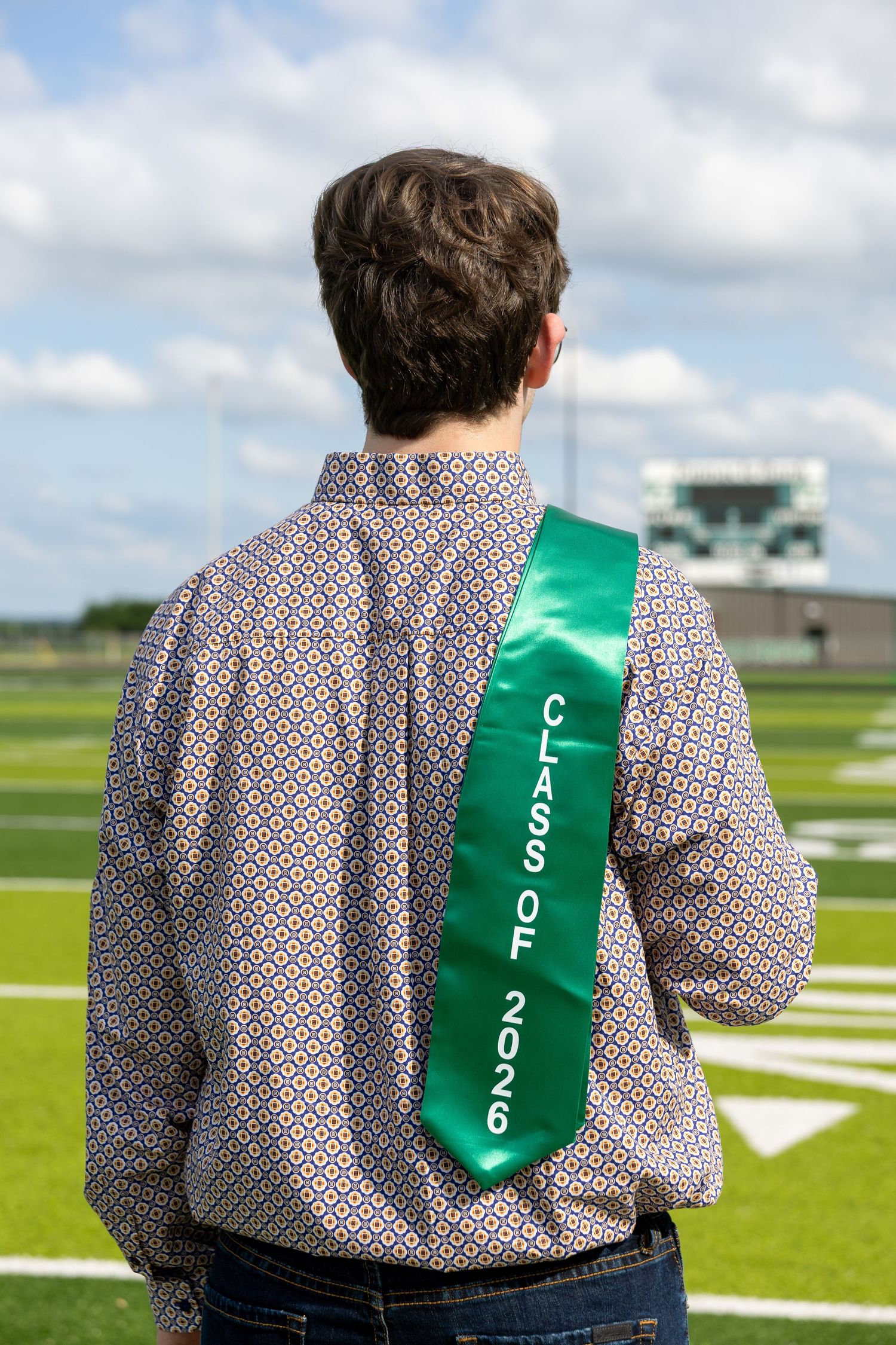Graduate wearing green Class of 2026 sash on patterned shirt standing on a football field from behind.