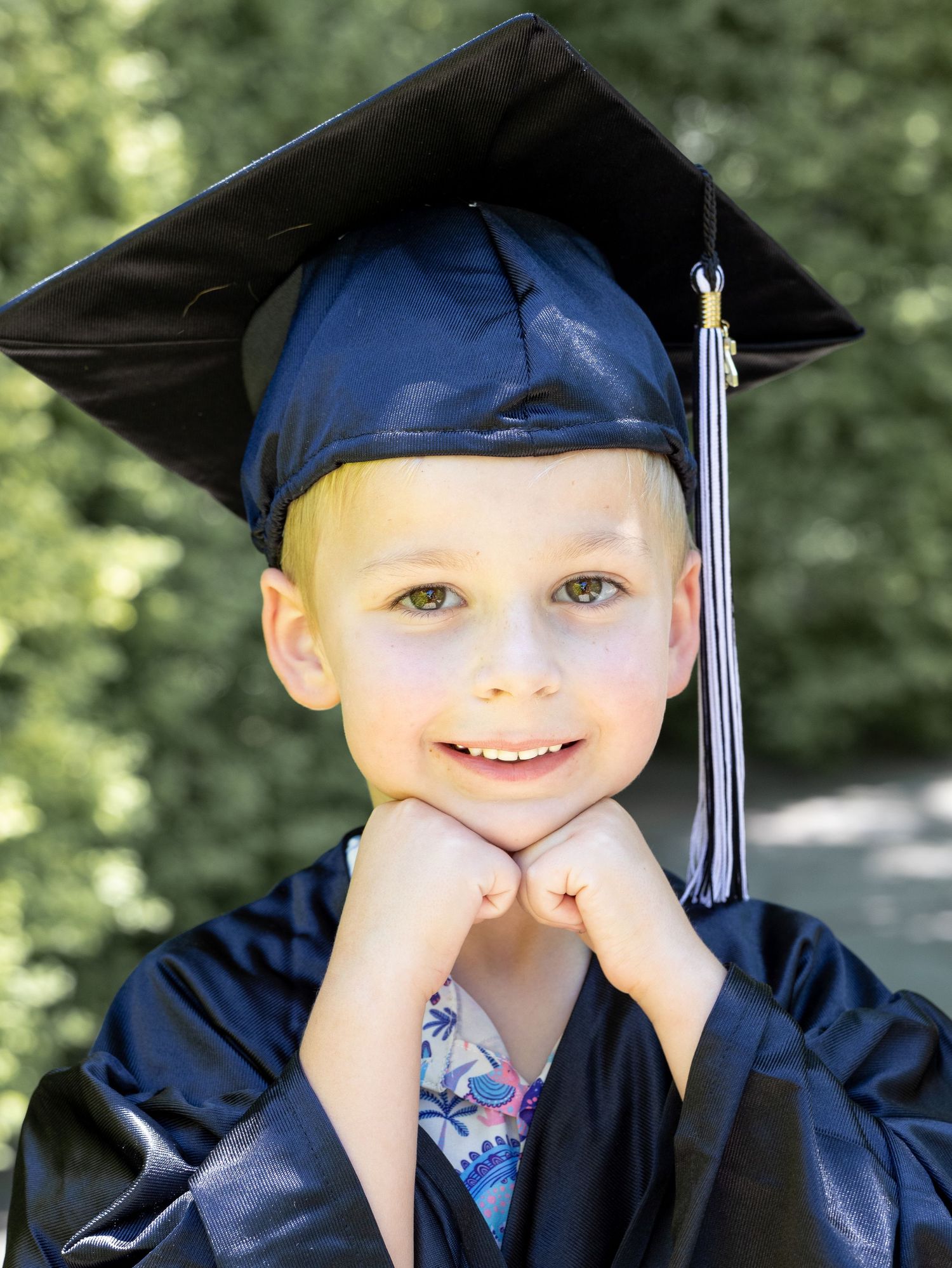 Young boy smiling in navy graduation cap and gown with gold tassel, resting chin on hands outdoors.