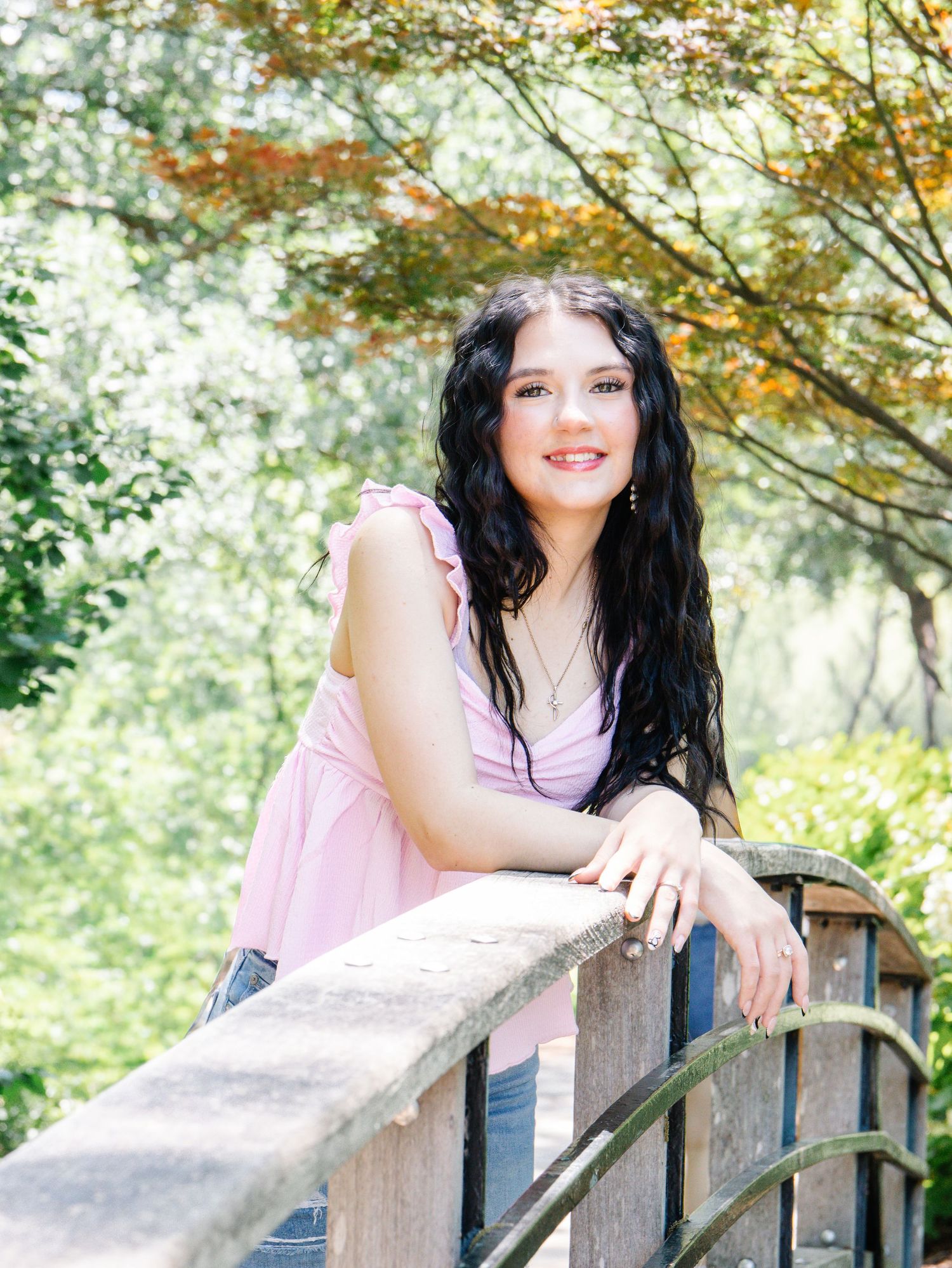 A smiling young woman with dark hair leans on a wooden railing surrounded by lush green trees.