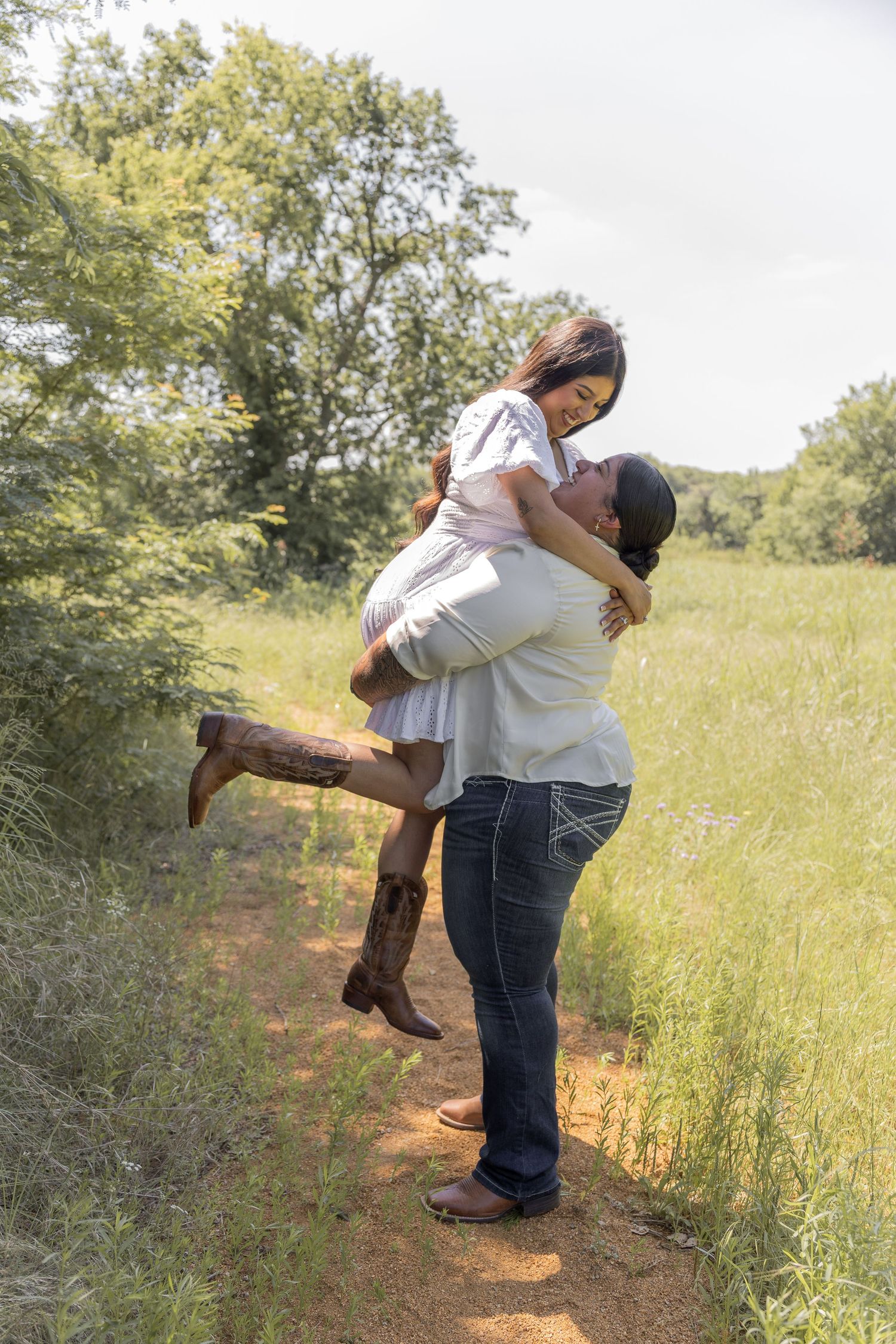 A man lifts and embraces a woman in a white dress and cowboy boots on a scenic green field path.