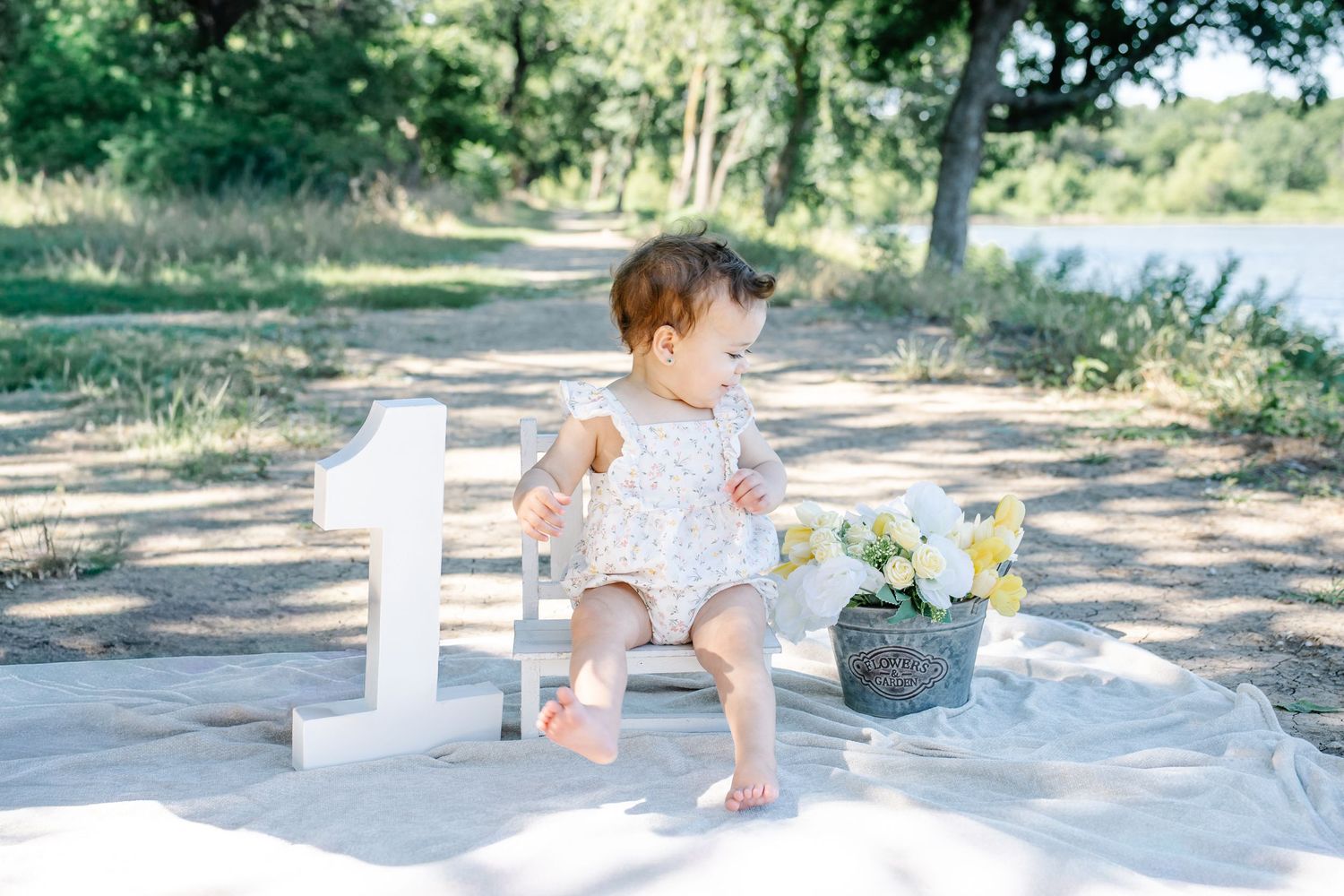 Baby in a white romper sits on a blanket beside a large white number one during an outdoor first birthday photoshoot. Saginaw Tx