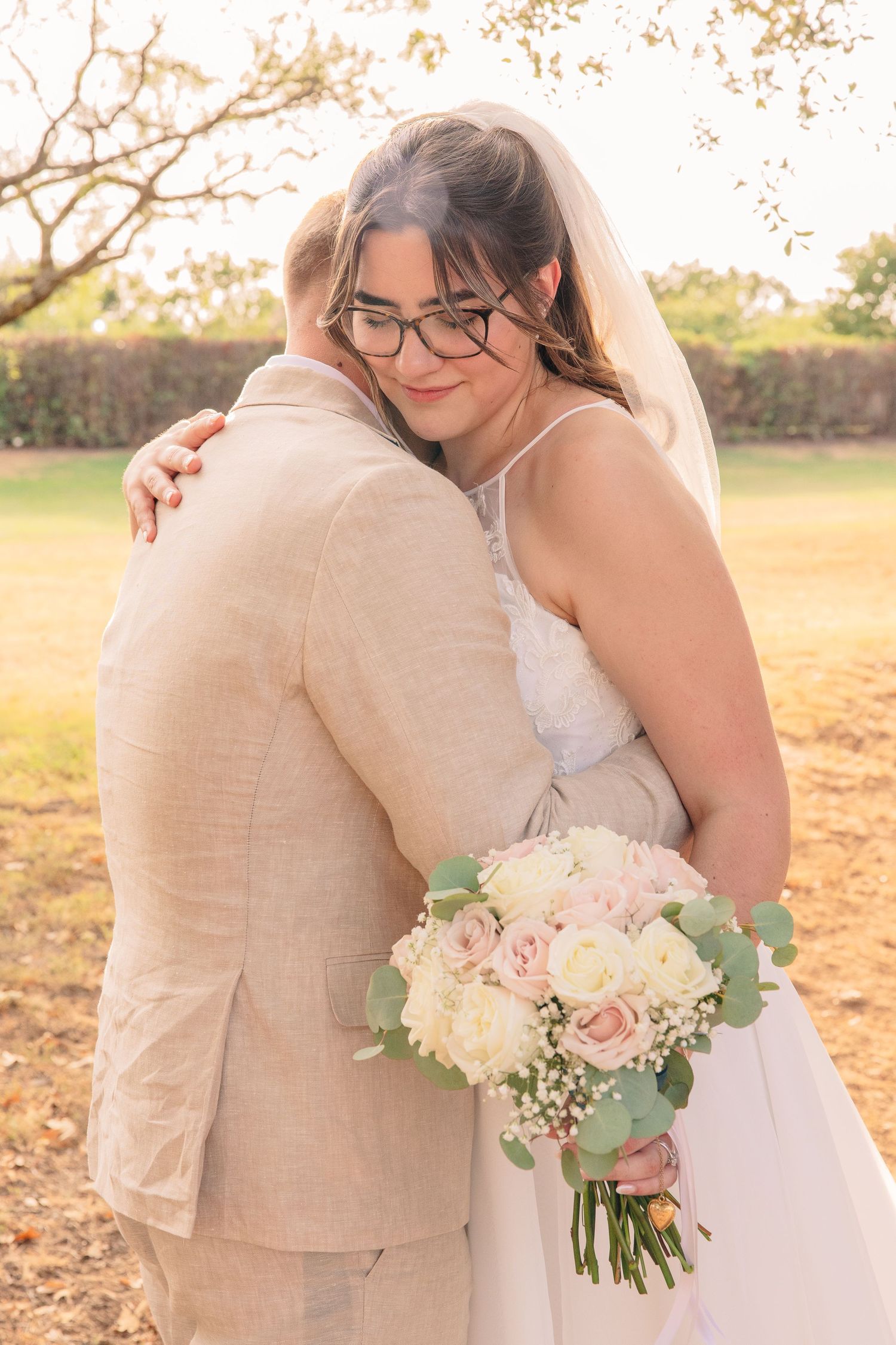 Bride in a white gown and veil holding a pink and white rose bouquet embraces a groom in a tan linen suit in a sunlit outdoor field.