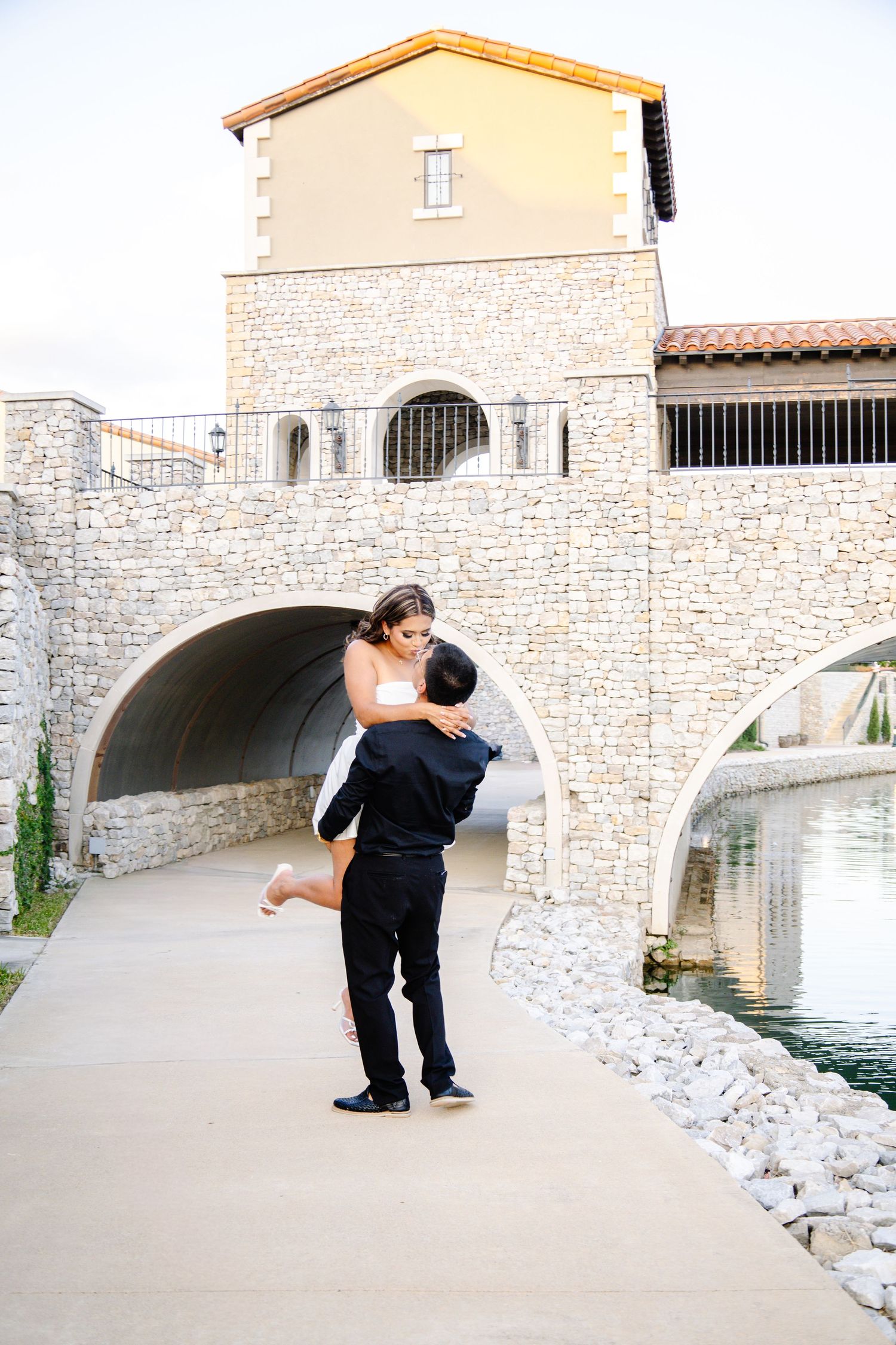 Couple embracing and lifting near a stone bridge and waterway at an outdoor venue during a romantic engagement photo session.
