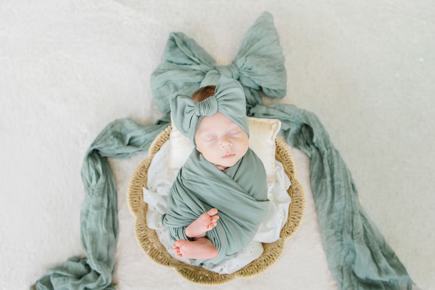 Newborn baby sleeping peacefully wrapped in a sage green swaddle with a matching bow headband in a wicker basket.