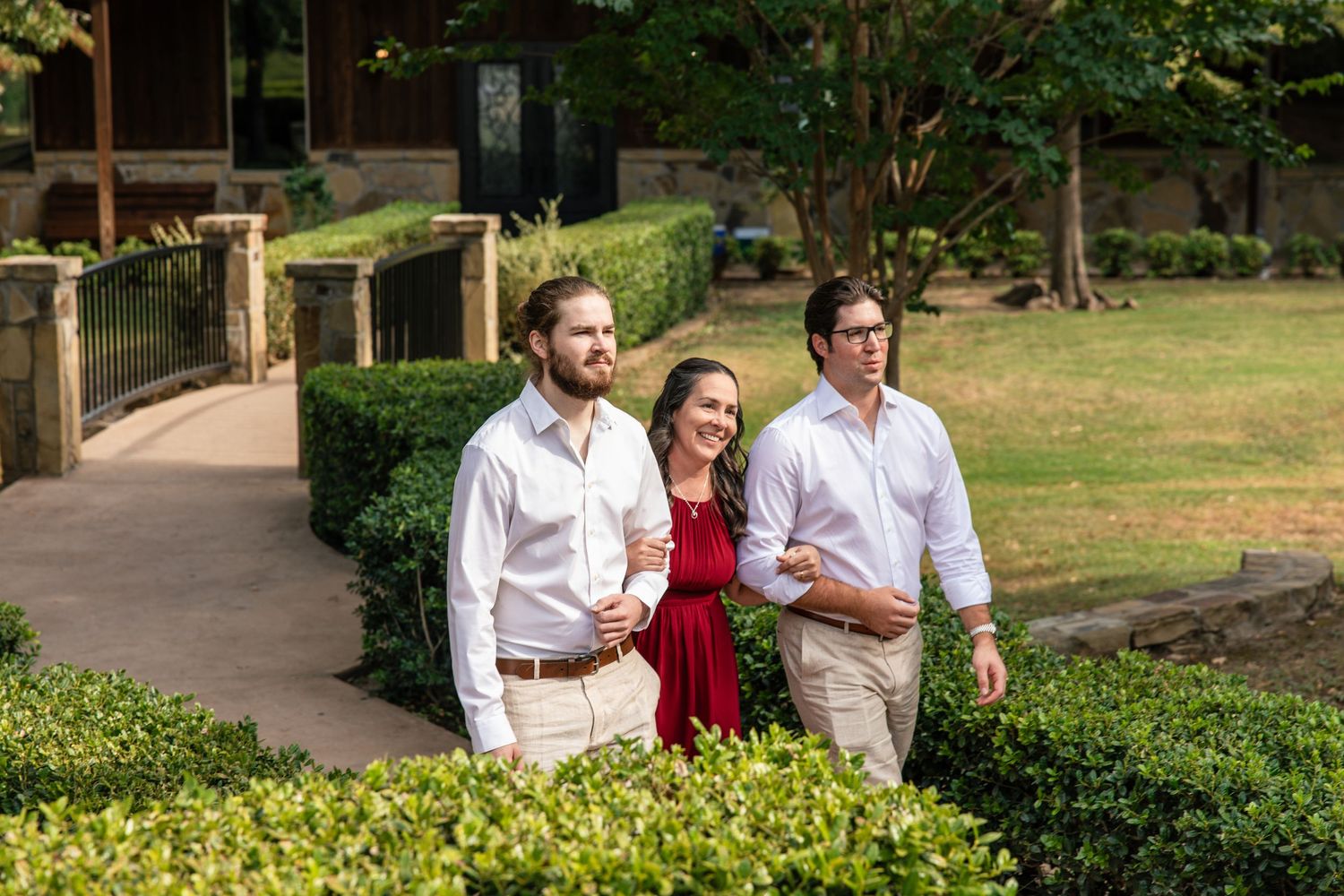 Two men in white button-up shirts and a smiling woman in a red dress walk together along a garden path lined with green hedges.
