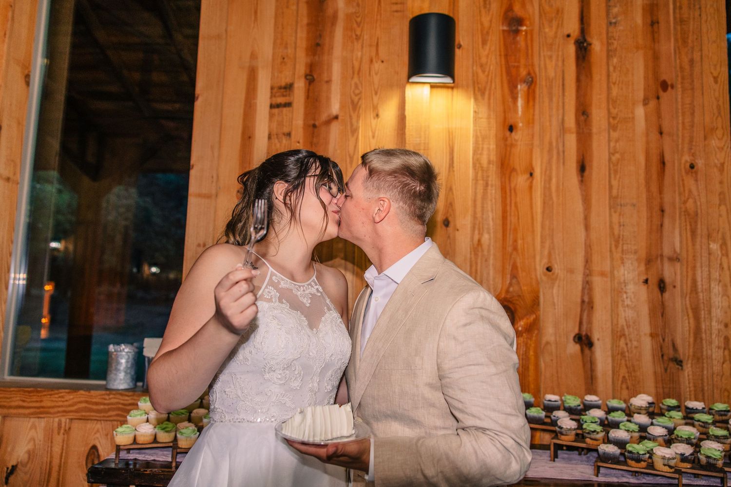 Bride and groom share a kiss while holding a slice of wedding cake beside a rustic wooden wall and a table of cupcakes.