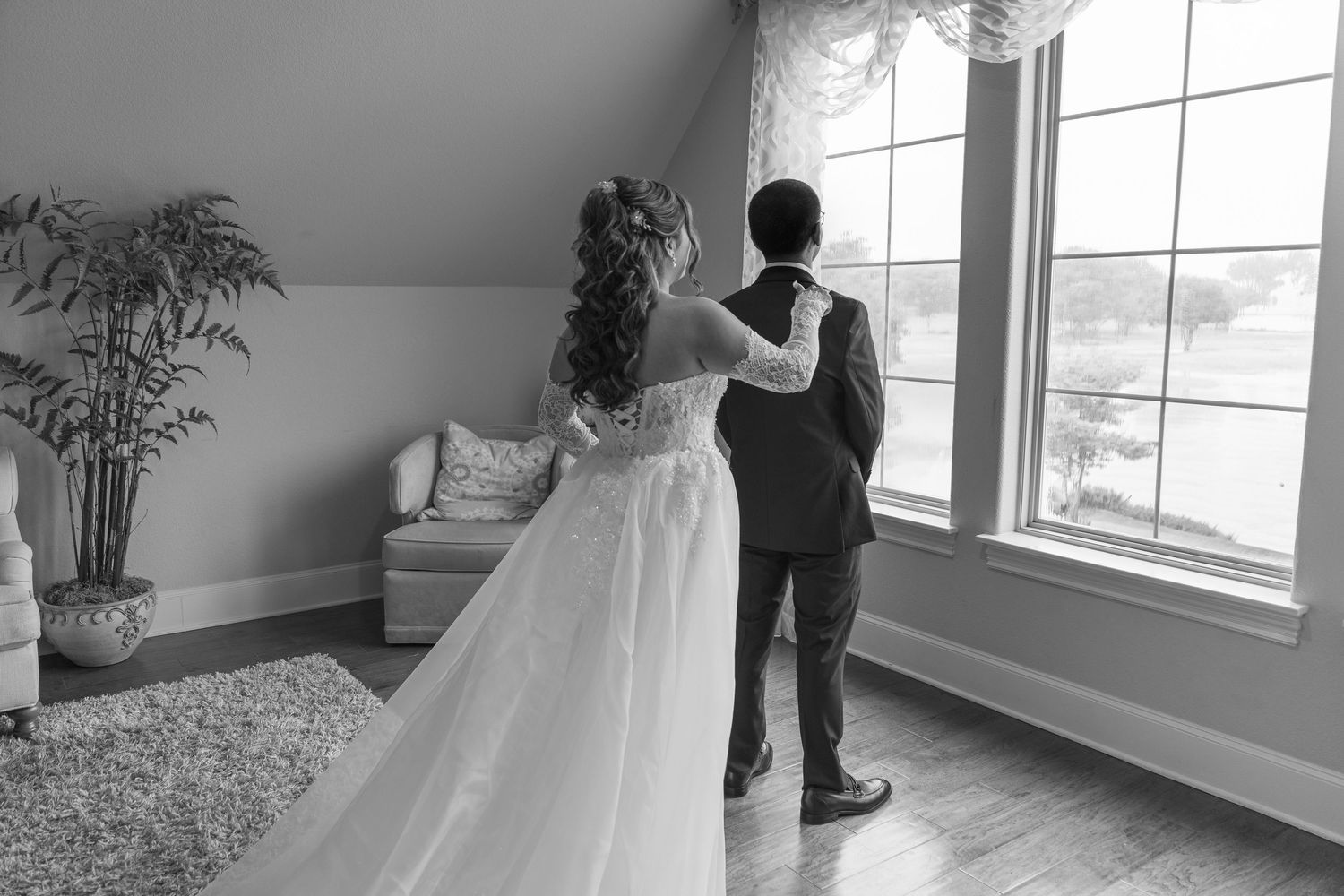 Black and white photo of a bride and groom standing together looking out a large window on their wedding day.