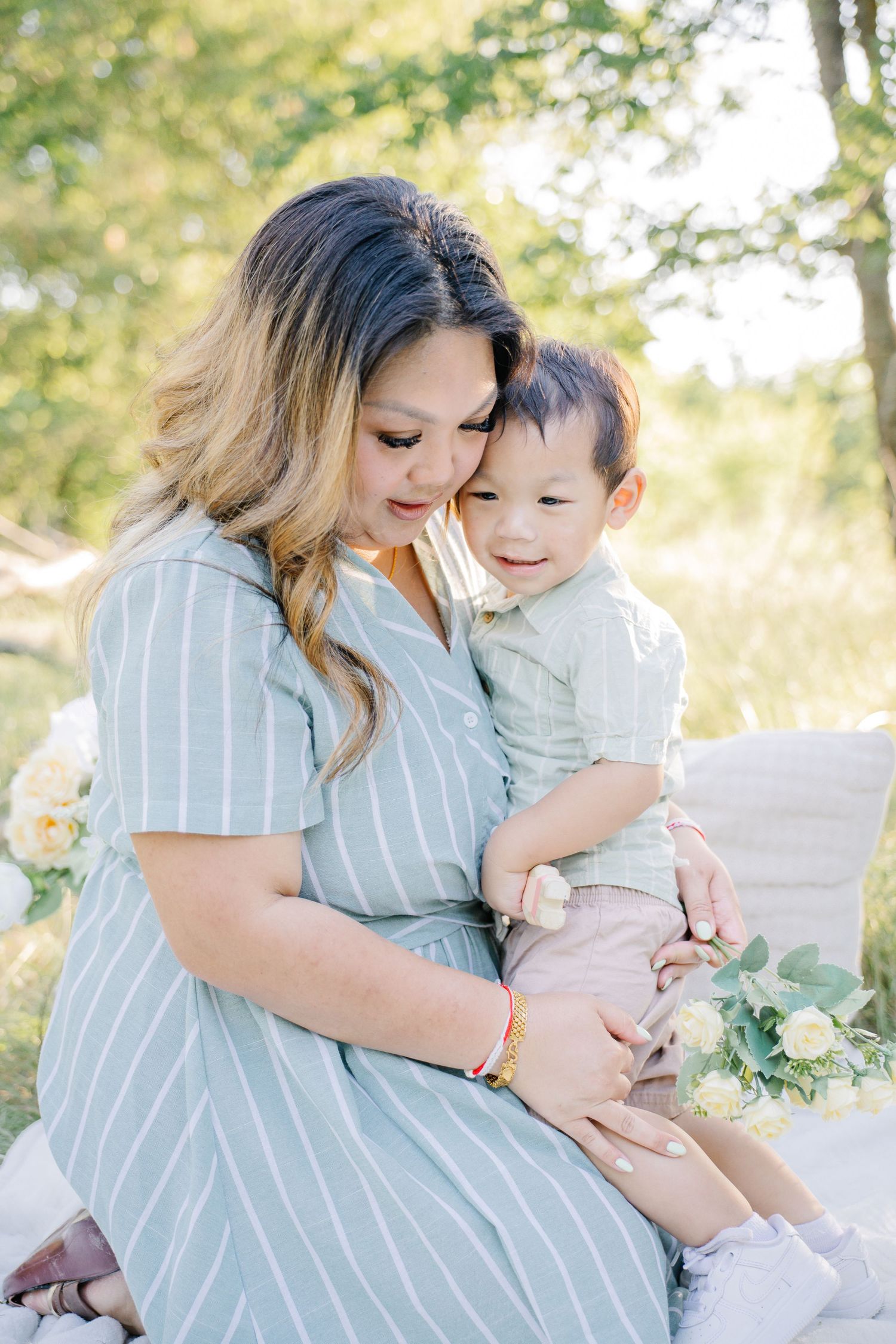 A mother in a striped dress tenderly holds her smiling toddler son outdoors during a family photo session.