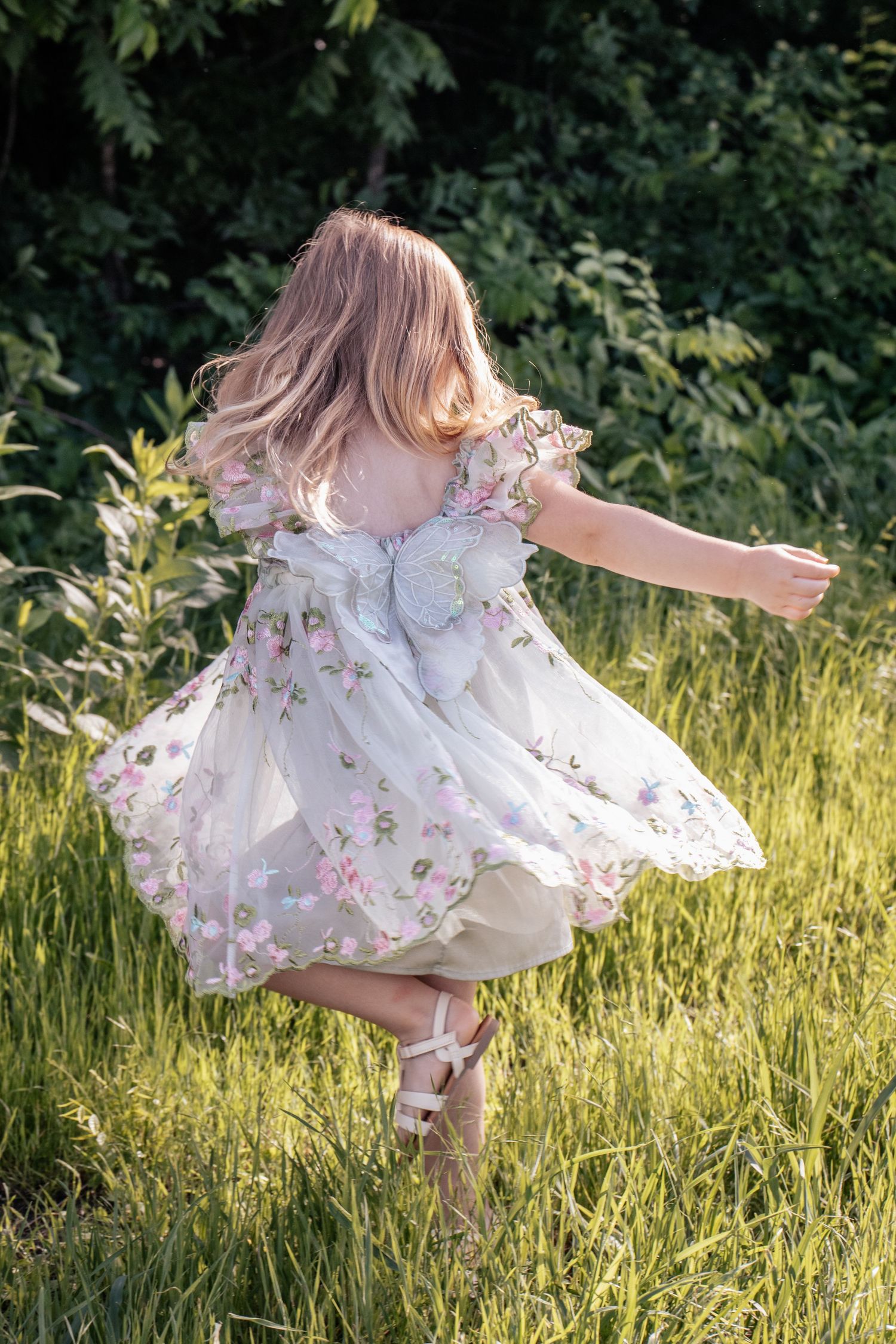 Young girl in floral dress with butterfly wings twirling in green meadow, captured from behind in natural sunlight.