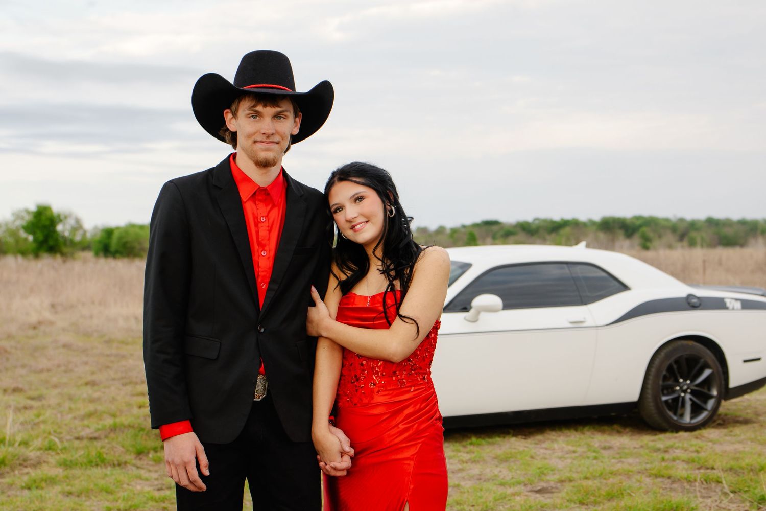 Couple in formal prom attire posing together in a field with a white Dodge Challenger.