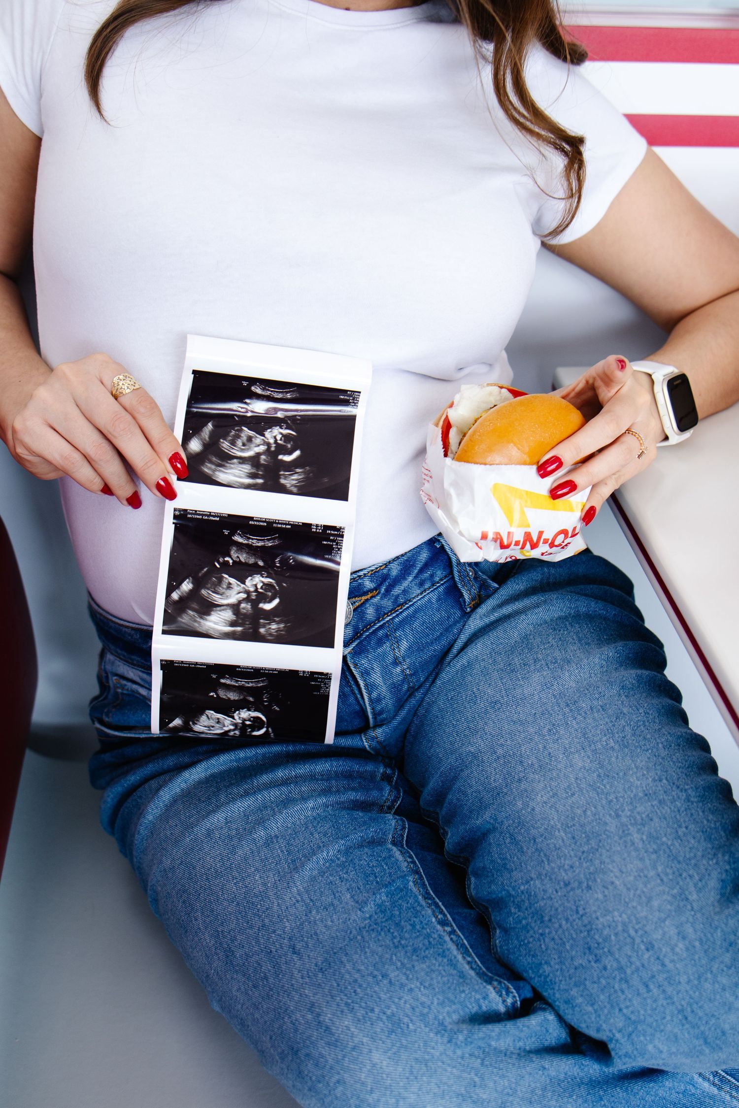 Pregnant woman holding ultrasound photos and an In-N-Out burger, showing her baby bump in a restaurant booth.