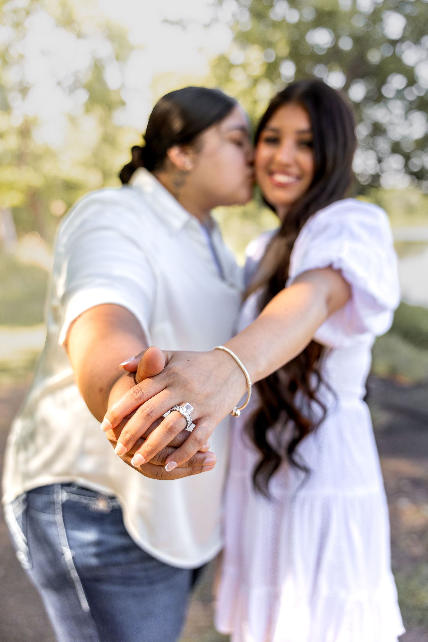 Couple showing engagement ring to camera outdoors, woman smiling in white dress while partner kisses her cheek.