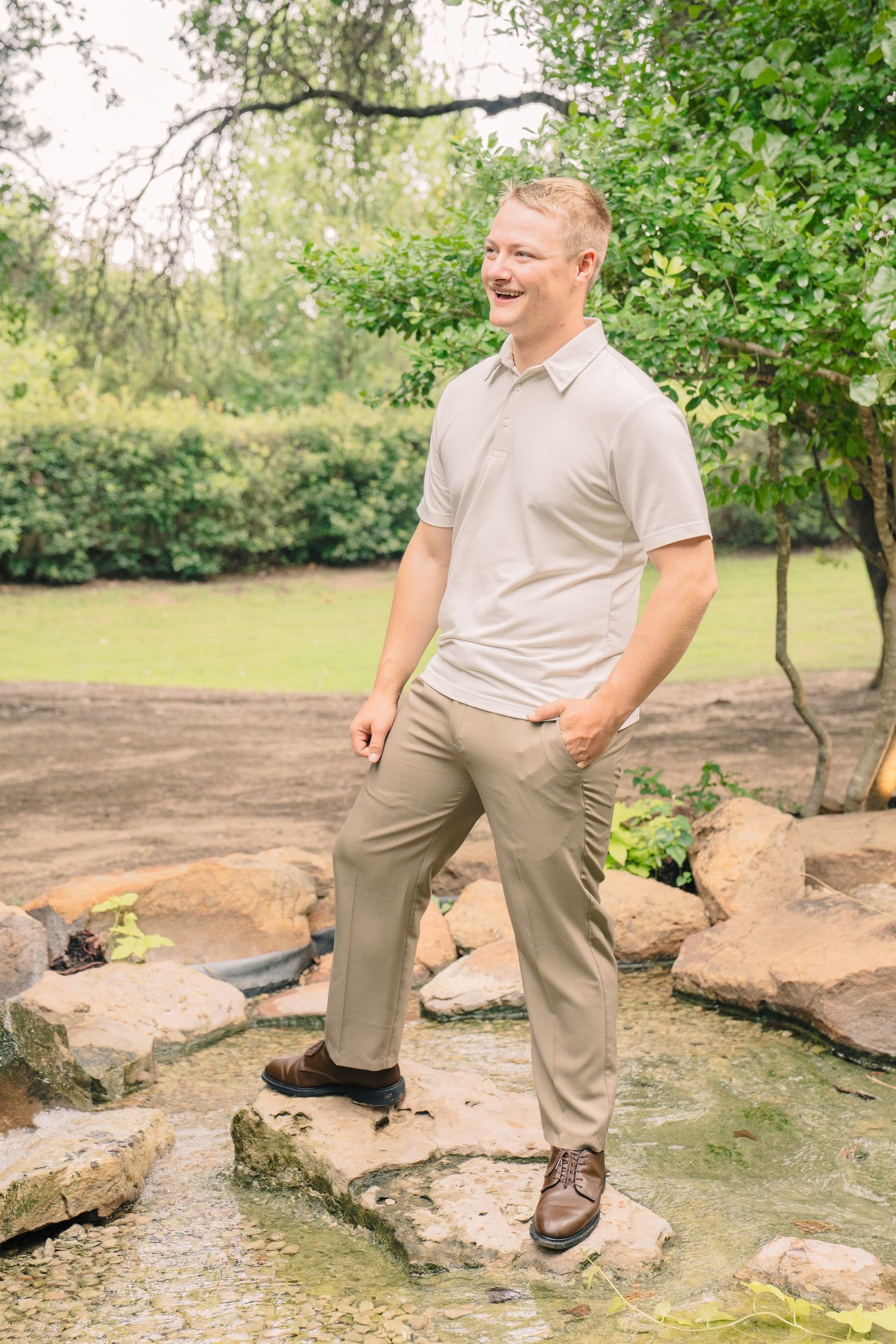 A smiling man in a beige polo shirt and khaki pants stands on rocks near a pond in a lush green garden.