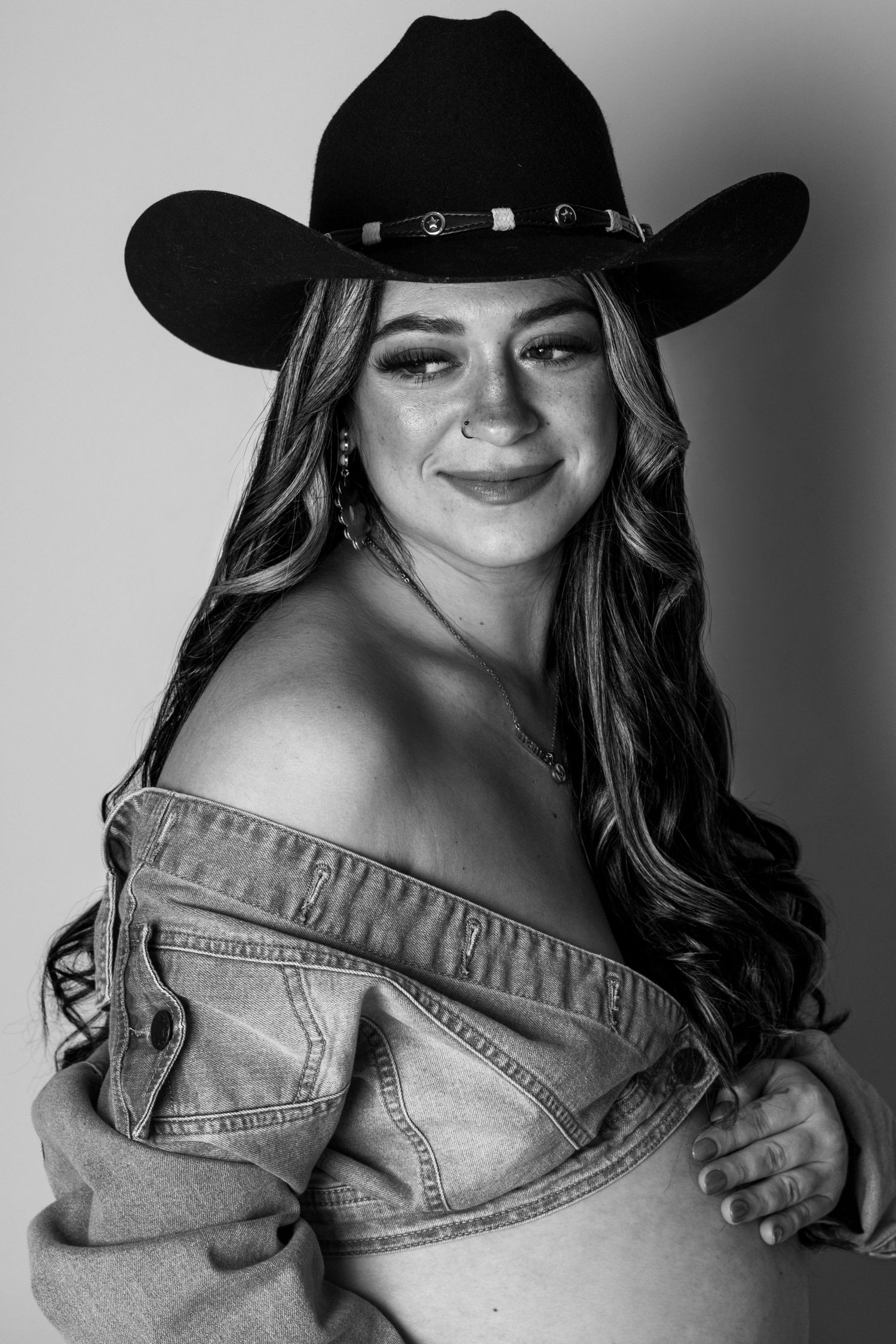Smiling pregnant woman in black cowboy hat and off-shoulder denim jacket, black and white maternity photo.