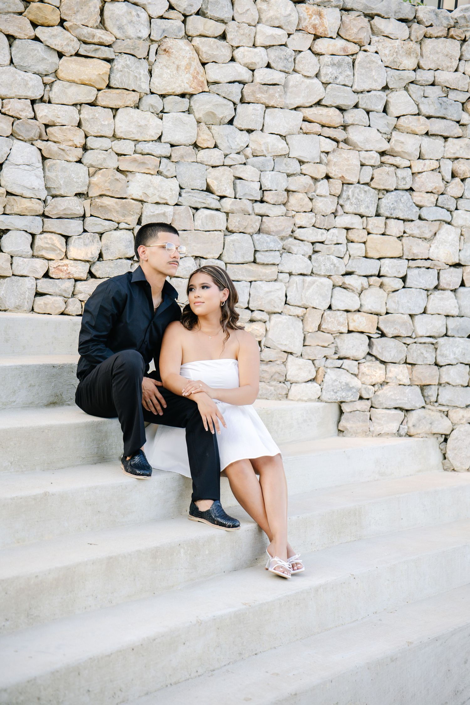 Couple sitting on stone steps embracing against a rustic stone wall backdrop.