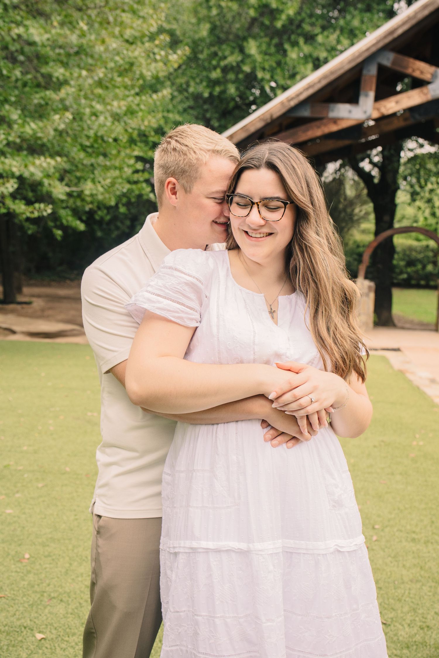 Man embracing woman in white dress from behind, sharing a tender moment during an outdoor engagement photography session.