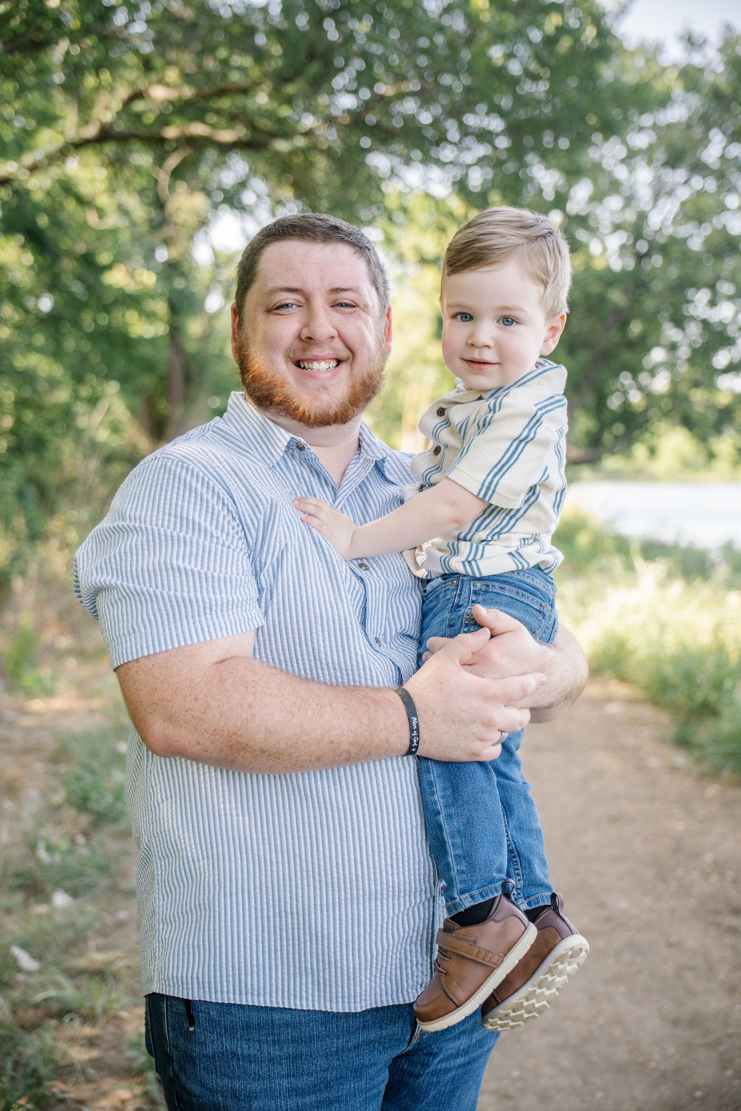Smiling bearded man in a blue striped shirt holds a toddler in a striped shirt and jeans on a sunny tree-lined outdoor path. Haslet tx